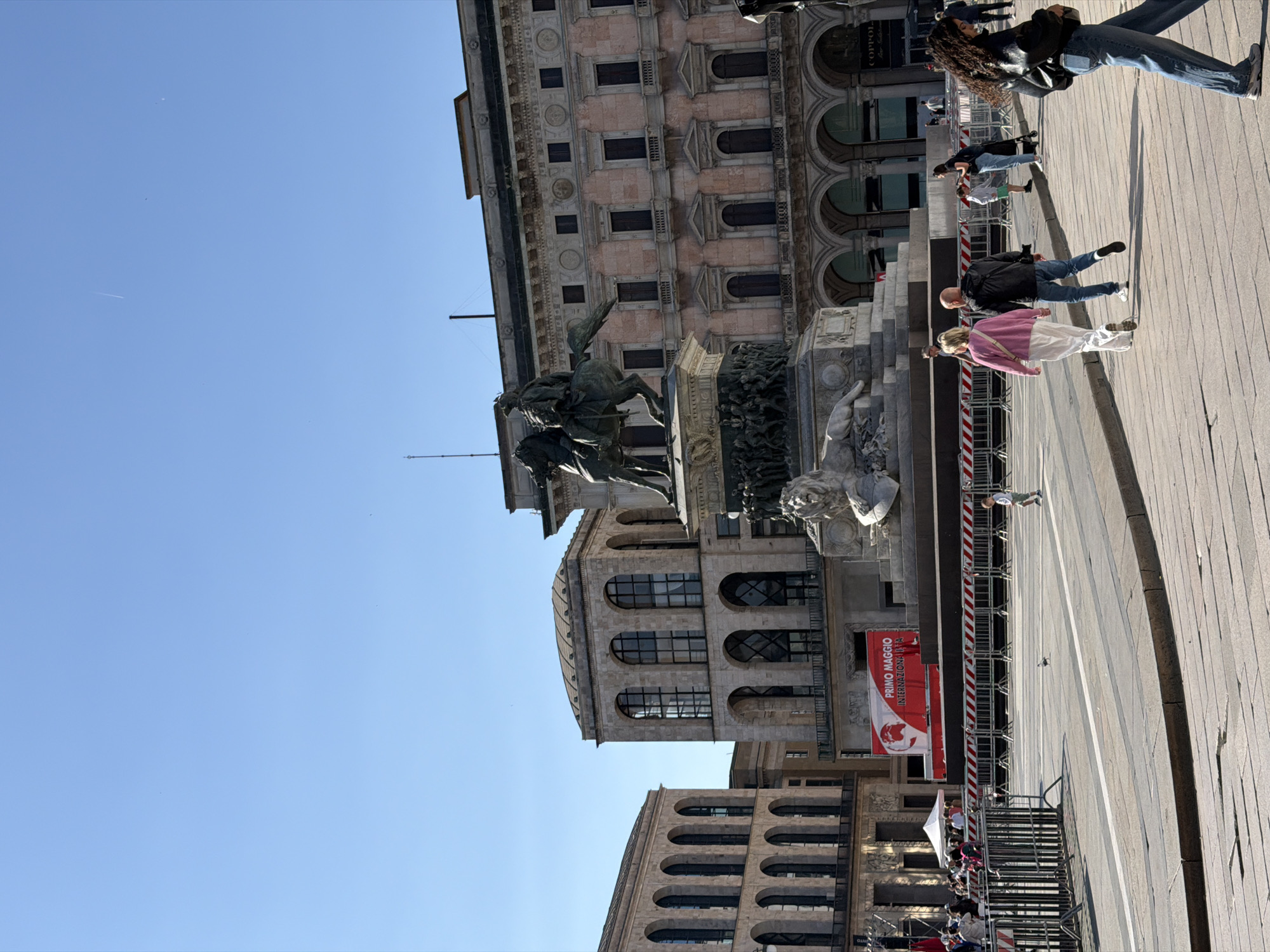 The image shows a view of the Piazza del Duomo in Milan, Italy. A prominent equestrian statue is centered in the frame, with ornate buildings in the background. People are walking and standing around the statue and in the plaza. A large red banner with white writing is visible to the right of the statue. The sky is clear and blue.