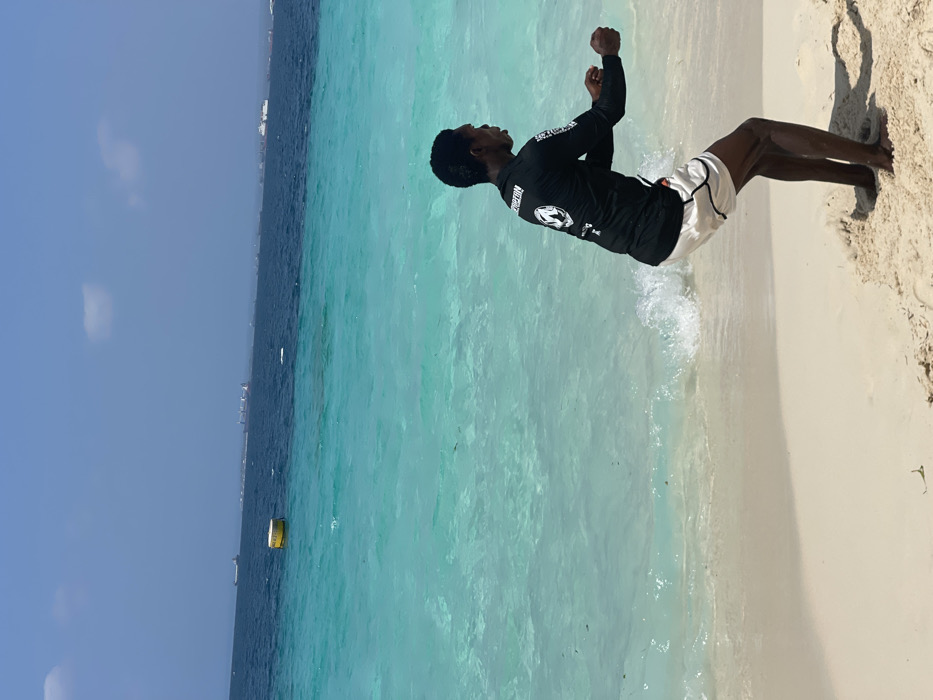 The image displays a sandy beach scene in Zanzibar, Tanzania, under a clear blue sky with minimal cloud cover. In the foreground, a male individual with a dark complexion is depicted in a crouched position, facing towards the right side of the frame. He is wearing a black long-sleeved rash guard with a white circular logo on the upper back, and light-colored shorts with a dark trim. His arms are extended forward with loosely clenched fists. He stands barefoot on the wet sand where small waves are breaking. The ocean water is a vibrant turquoise near the shore, transitioning to a deeper blue further out. A yellow cylindrical buoy floats in the midground water on the left. Several large cargo or passenger vessels are visible on the distant horizon, indicative of maritime activity.