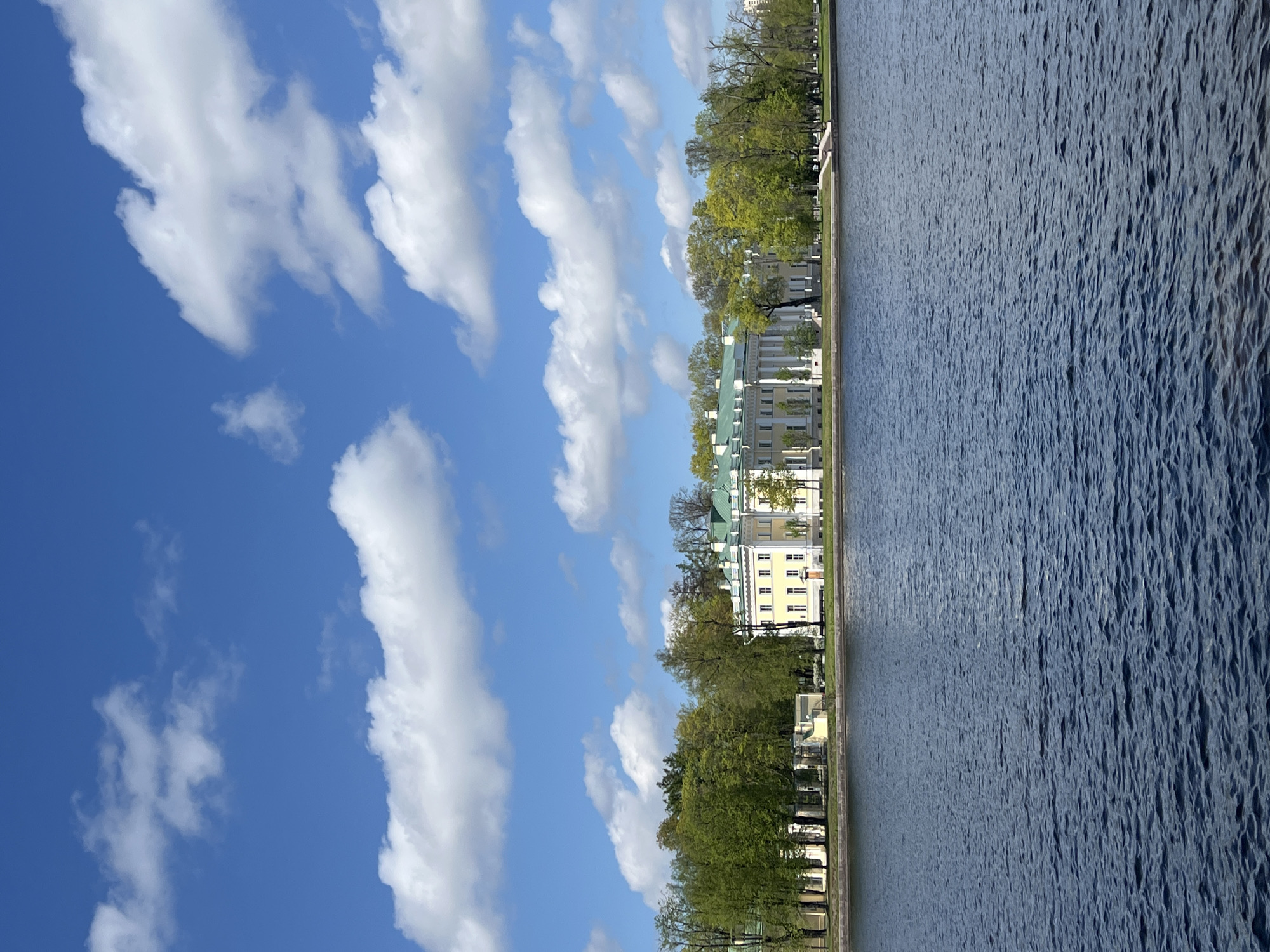 The image shows a view along the Neva River in Saint Petersburg, Russia. On the right, the Neva's dark, rippled water fills the scene. Adjacent to the water, on the left, there is a line of trees, and behind the trees is a yellow and white building with a green roof. To the left of this area, a clear blue sky is filled with bands of white clouds.