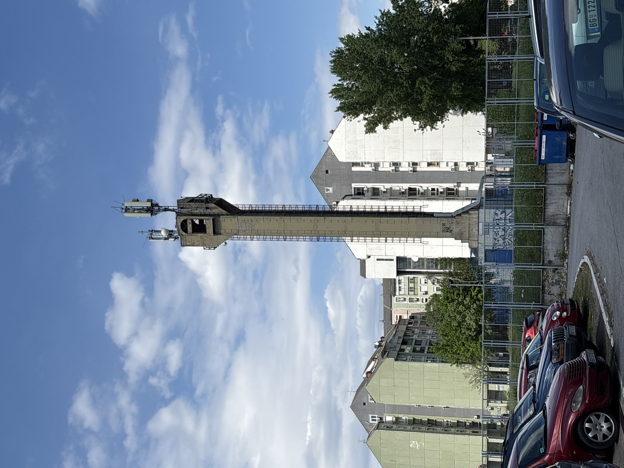 The image shows the Žrnovo transmitter tower in Belgrade, Serbia. The tower is a tall, concrete structure with antennas at the top. It is located next to a building and a parking lot with several cars. The sky is blue with clouds.