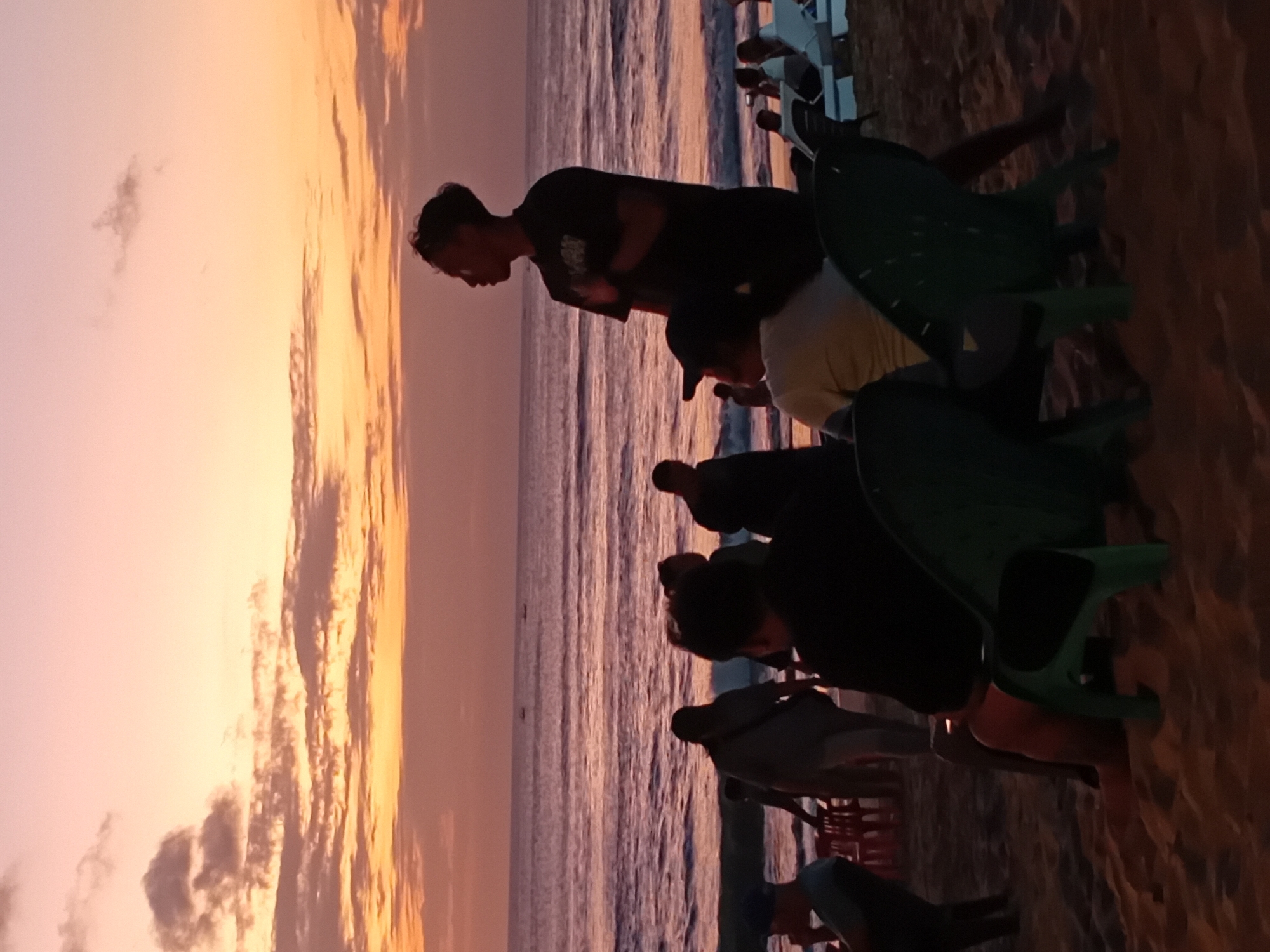 A group of people are on a beach in Kuta, Indonesia, at sunset. The sky is a gradient of pink and orange, with scattered clouds. The ocean is calm, with gentle waves. Several people are visible on the sand, some sitting in chairs and others standing. In the foreground, two green plastic chairs are empty. A man in a black t-shirt stands in silhouette, looking out at the ocean. Behind him, other figures are indistinct in the dim light. Some lounge chairs are visible further down the beach, with more people sitting on them. The sand is a warm, golden color.