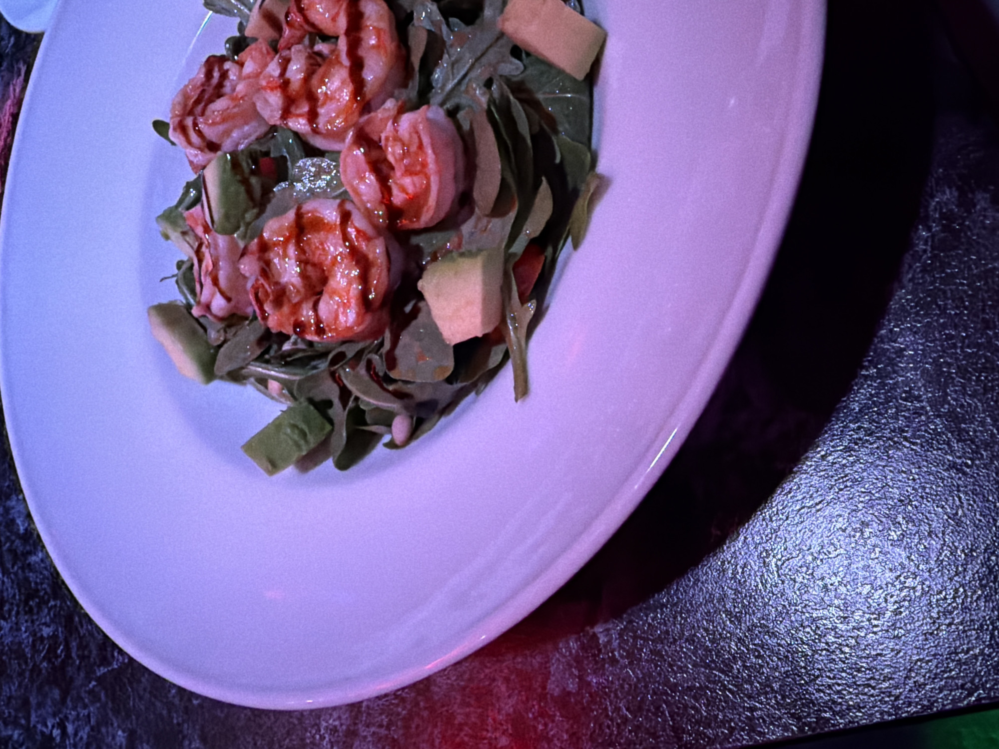 A shrimp and avocado salad is presented on a white, slightly oval plate. The plate is sitting on a dark textured surface. The image is cast in a purple-blue hue. No people or other objects are visible. The location is unspecified; Chaikovsky, Russia, is not inferable from the image.