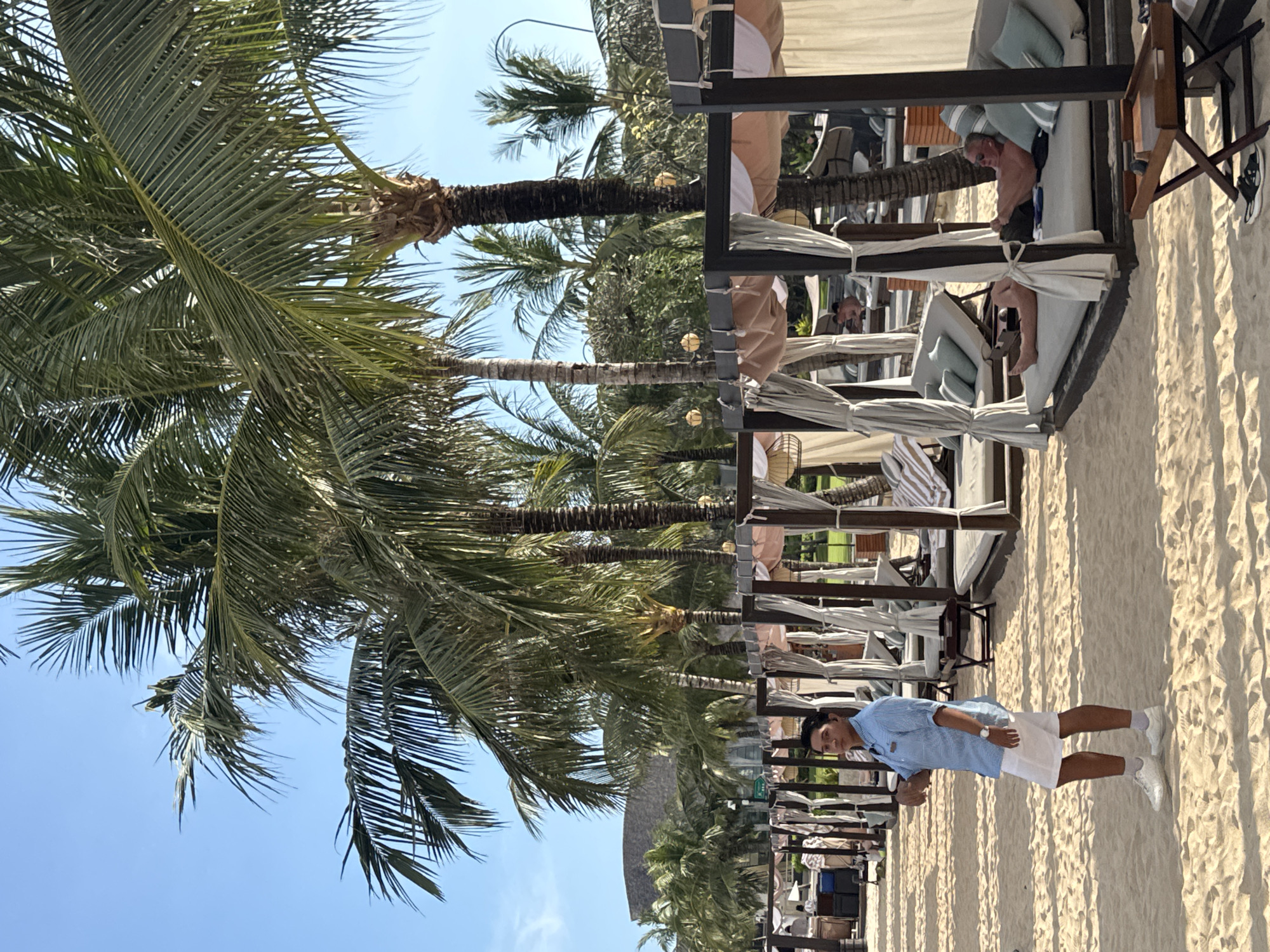 A full shot captures a man in a short-sleeved light blue shirt and white shorts, standing on a sandy beach in Kuta Selatan, Indonesia. The beach is lined with cabanas, some of which are occupied by people lounging. Palm trees stretch into a clear blue sky, creating a tropical backdrop.