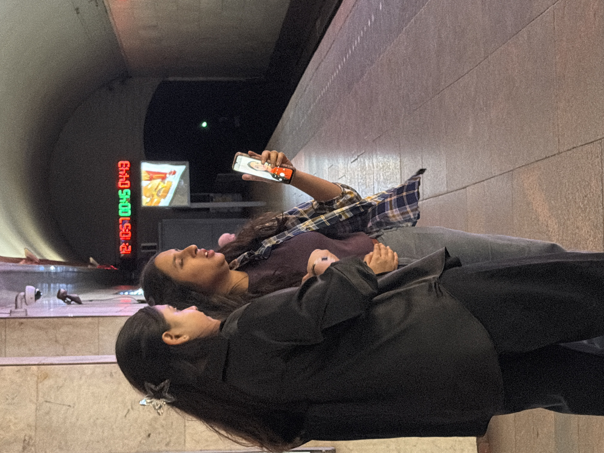 Two women are standing on a subway platform, possibly in Tbilisi, Georgia, looking at a smartphone screen held by the woman on the right. The woman on the left is wearing a black jacket and dark pants, and has a silver star-shaped hair clip. The woman on the right is wearing a purple shirt and a plaid shirt over it, and gray pants. The smartphone displays a video call with a woman's face on the screen. In the background, a digital clock shows the time "12:05:45" in red and green digits. The platform is made of tiled concrete, and the subway tunnel curves into the distance. There are safety markings on the edge of the platform.