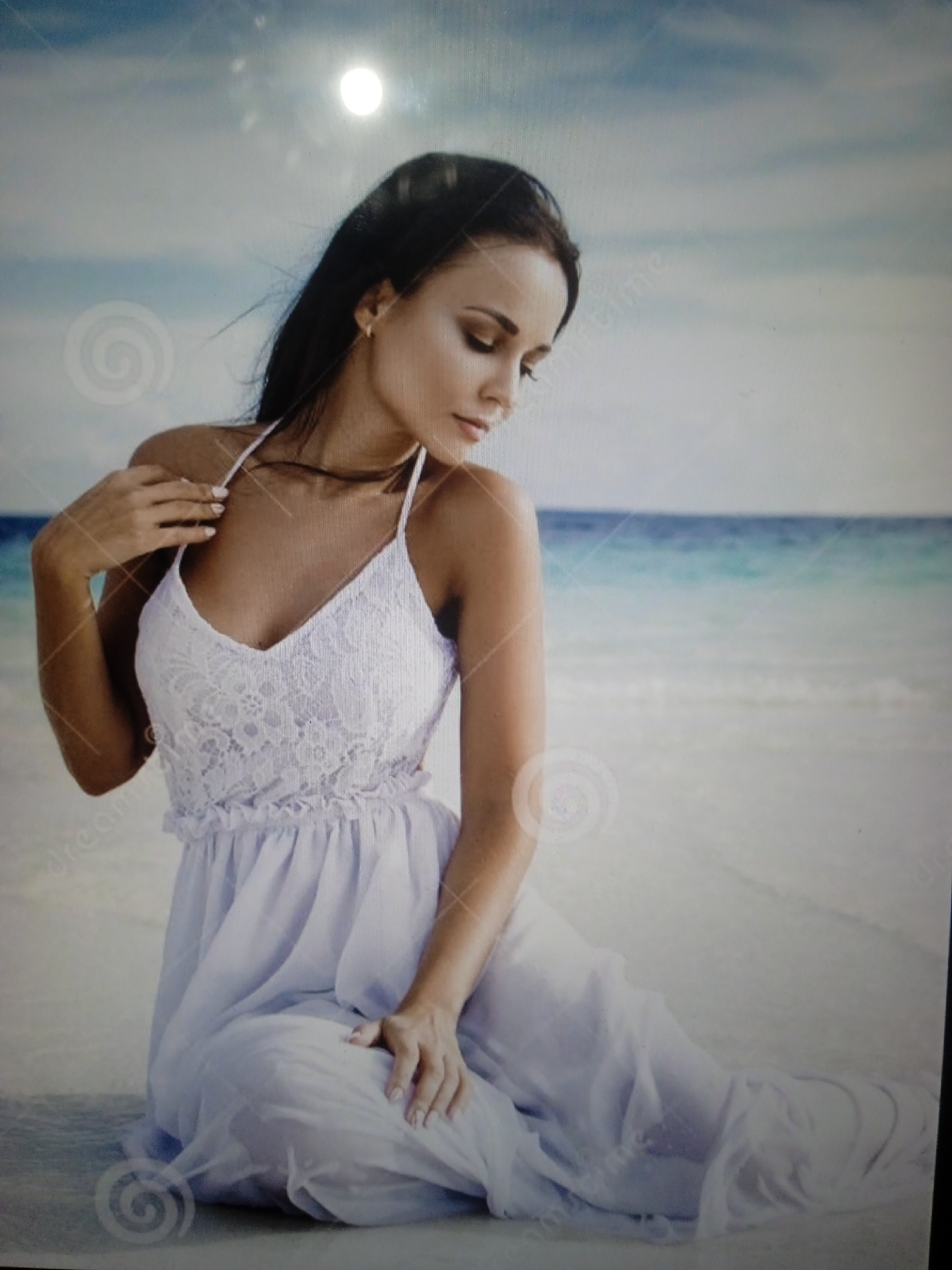 A woman in a white lace-trimmed dress sits on a sandy beach. The ocean is visible in the background, with a slightly cloudy sky above. The sun appears as a bright, blurred orb in the sky. The woman has dark hair and is looking downwards. Her hands are resting on her lap and her chest. The dress is long and flowing. The scene is serene and appears to be a professional photo shoot, likely with watermarks present. There is no indication of Chernivtsi, Ukraine.