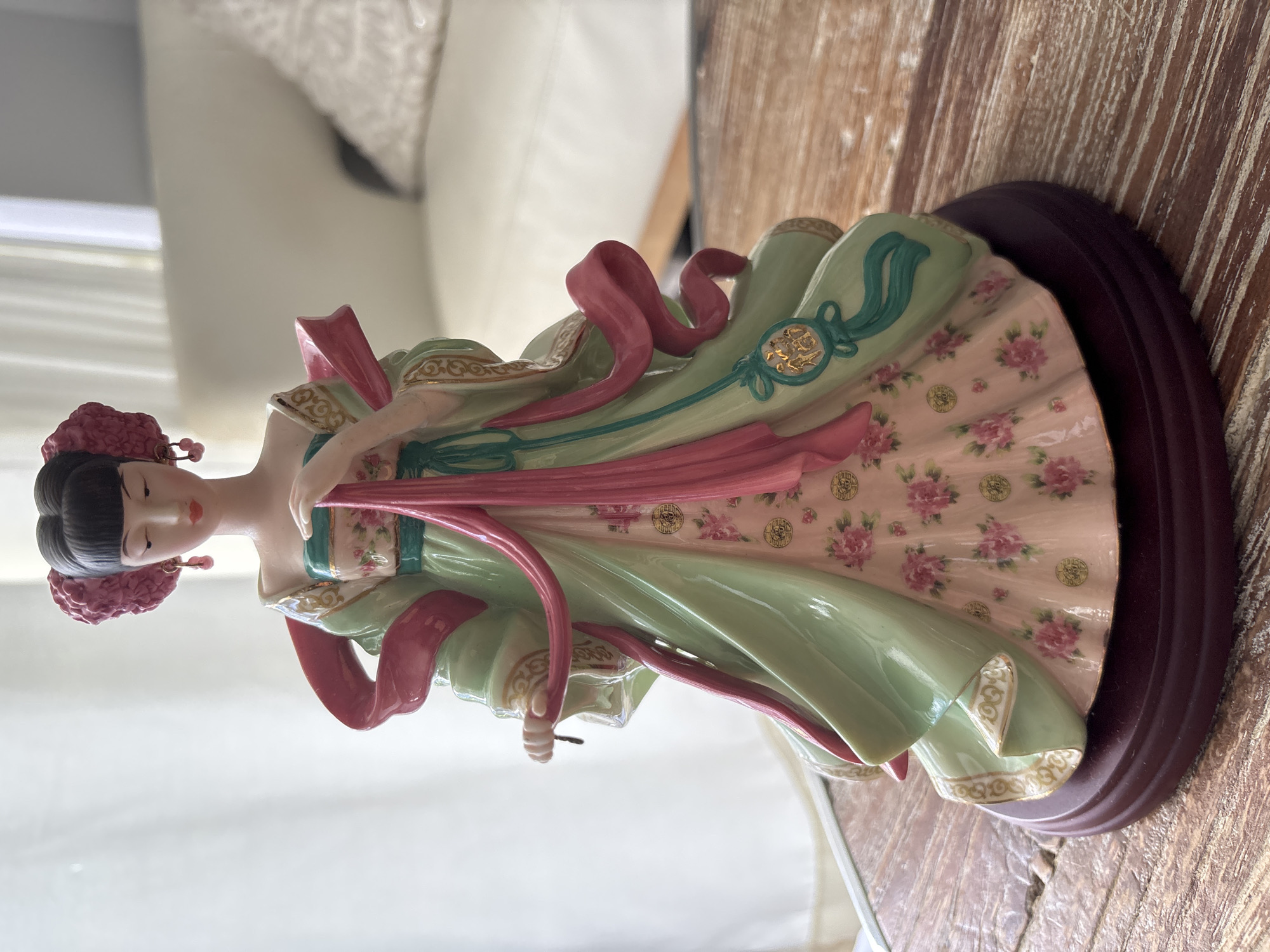 A porcelain figurine of an Asian woman stands on a wooden table. The figurine is dressed in a green robe with pink accents and floral patterns. The woman has dark hair styled in an updo with a pink accent. She is wearing dangling earrings and has her hands clasped in front of her. The figurine is on a dark red circular base. The background is blurred, showing a couch and a window with curtains. The scene is indoors, likely in a residential setting in Aventura, United States of America.