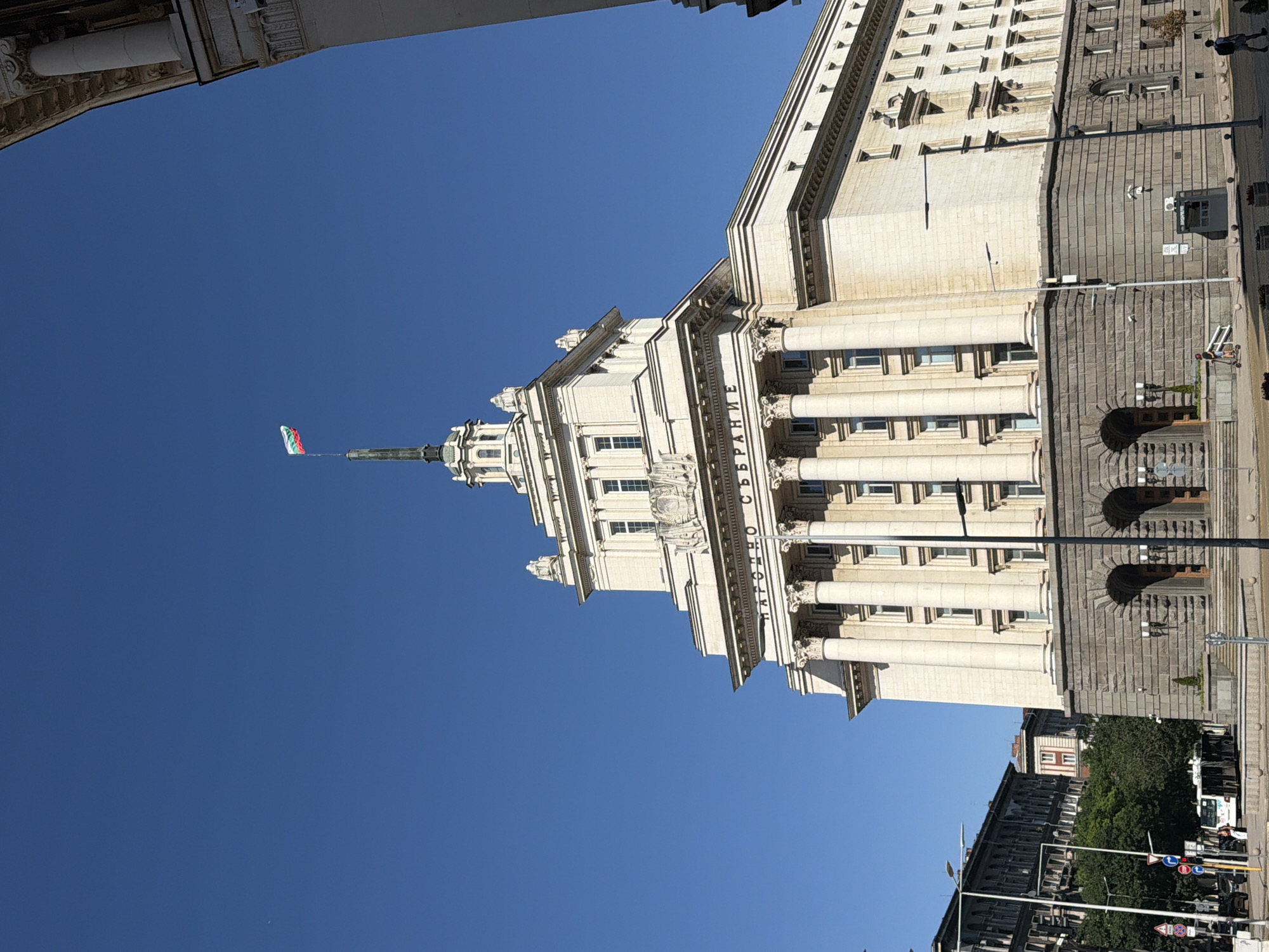 The image captures the National Assembly of Bulgaria, also known as the Narodno sabranie, situated in the capital city of Sofia. The grand building, constructed with light-colored stone, features a prominent facade with numerous columns and ornate architectural details. At the pinnacle of the building, the Bulgarian flag, a tricolor of white, green, and red, flies proudly against a clear blue sky.
In the foreground, a street scene unfolds. A modern white bus or trolleybus is visible on the left side, indicating public transportation is in operation. Several traffic signs and streetlights dot the area. Pedestrians are present, with one individual riding a scooter and another walking on the sidewalk. The street appears to be paved, and planters with small trees are arranged along the edge. The architecture of adjacent buildings, visible on the left, suggests an urban environment. The overall impression is a sunny day in a bustling city, with a focus on the historical and governmental significance of the building.