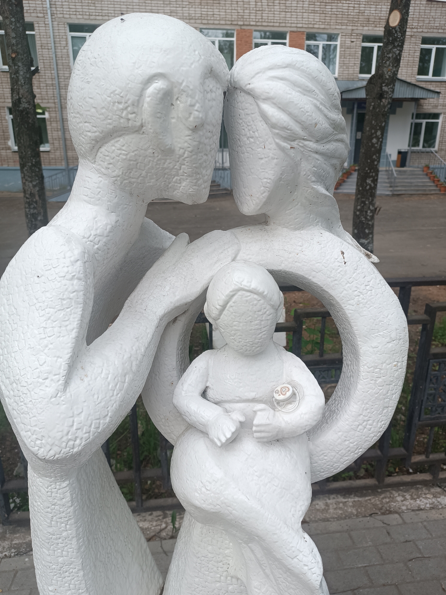 A white painted sculpture of a mother, father, and child stands outdoors in Chaikovsky, Russia. The father is on the left side of the sculpture, with his arm around the child and his head touching the mother's head. The mother is on the right side, also with her arm around the child. The child is in the center of the sculpture, holding a pacifier. The background consists of a grey building with white trim, a tree, a black fence and a stone sidewalk.