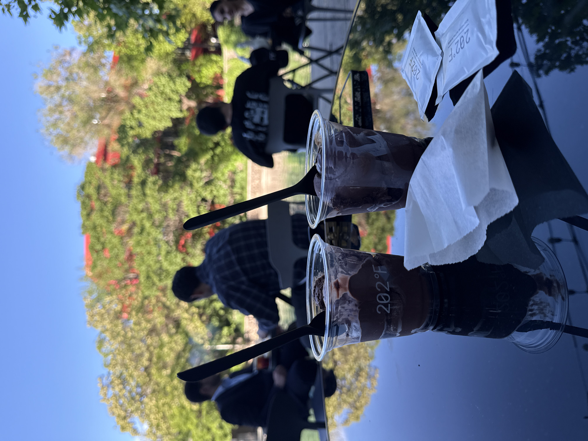 The image shows a table setting at an outdoor café, potentially in Yerevan, Armenia. Two clear plastic cups contain chocolate desserts, each with a black spoon inserted. Sugar packets and a white napkin are on the table. The reflective surface shows the silhouettes of people sitting at tables in the background, including individuals in casual attire. The background features trees and a blue sky. The cups are labeled with "202°F".