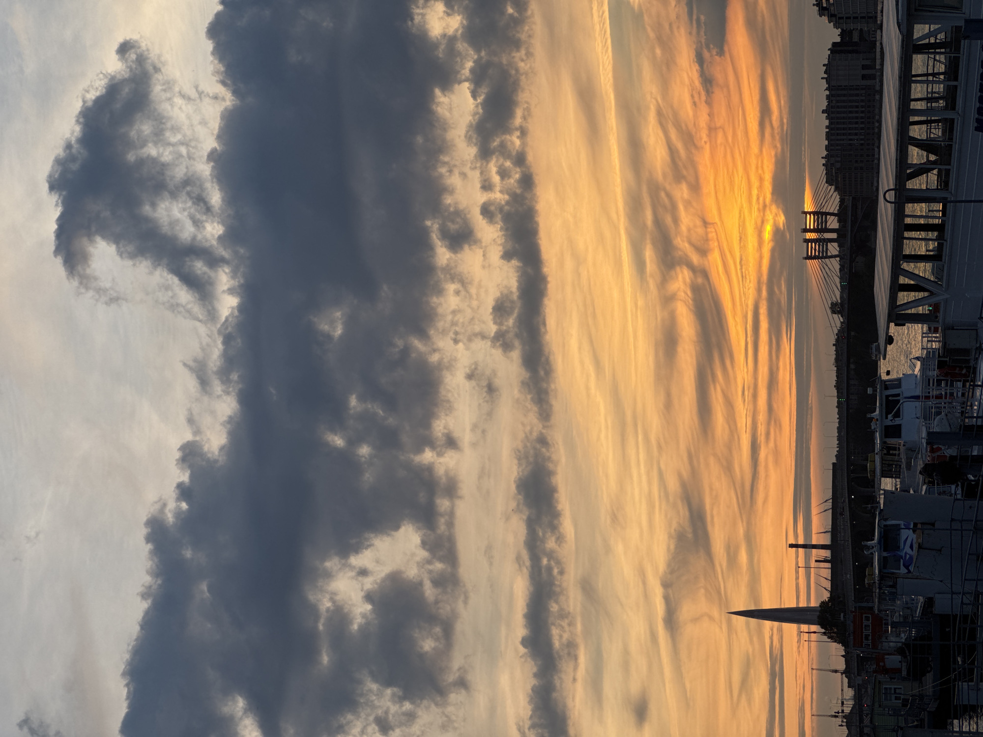 The image captures a sunset over the Neva River in Saint Petersburg, Russia. The sky is dominated by dramatic clouds, with dark gray formations in the upper portion transitioning to vibrant oranges and yellows closer to the horizon. The sun is partially obscured by clouds, casting a warm glow across the scene.
In the foreground, several boats are docked along the riverbank. These vessels appear to be for passenger transport, with some having covered seating areas. The architecture of Saint Petersburg is visible in the background, including a distinctive tall, spire-like building and bridges spanning the river. The overall atmosphere is one of twilight, with the city lights beginning to appear in the distance.