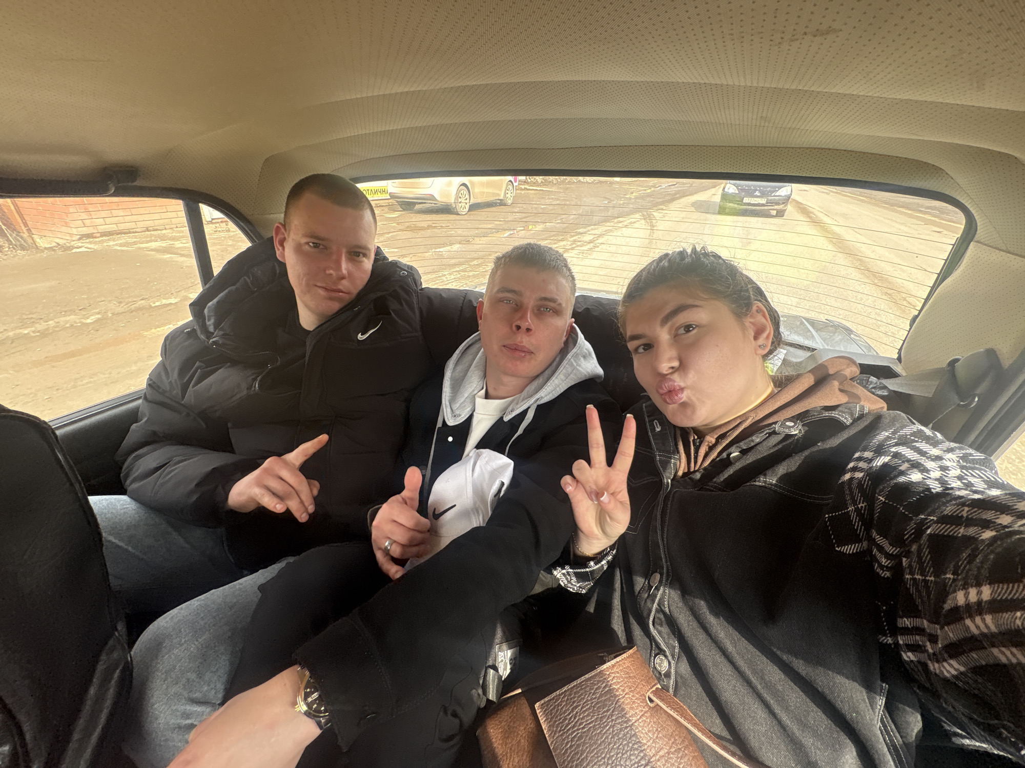 Three individuals are seated in the back of a car. The individual on the left is male, wearing a black puffer jacket, light blue jeans and is pointing to the right. The individual in the middle is also male, wearing a light grey hoodie and a dark jacket and is giving a thumbs up. The individual on the right is female, wearing a black denim jacket with plaid sleeves over a brown hoodie and is holding up two fingers. She is pursing her lips. A brown leather handbag is at the bottom of the frame. Outside the car, the scene suggests a town or city street in Chernushka, Russia. There are light-colored brick buildings and multiple cars, one of which is silver, parked in the distance. The road appears to be wet or muddy.