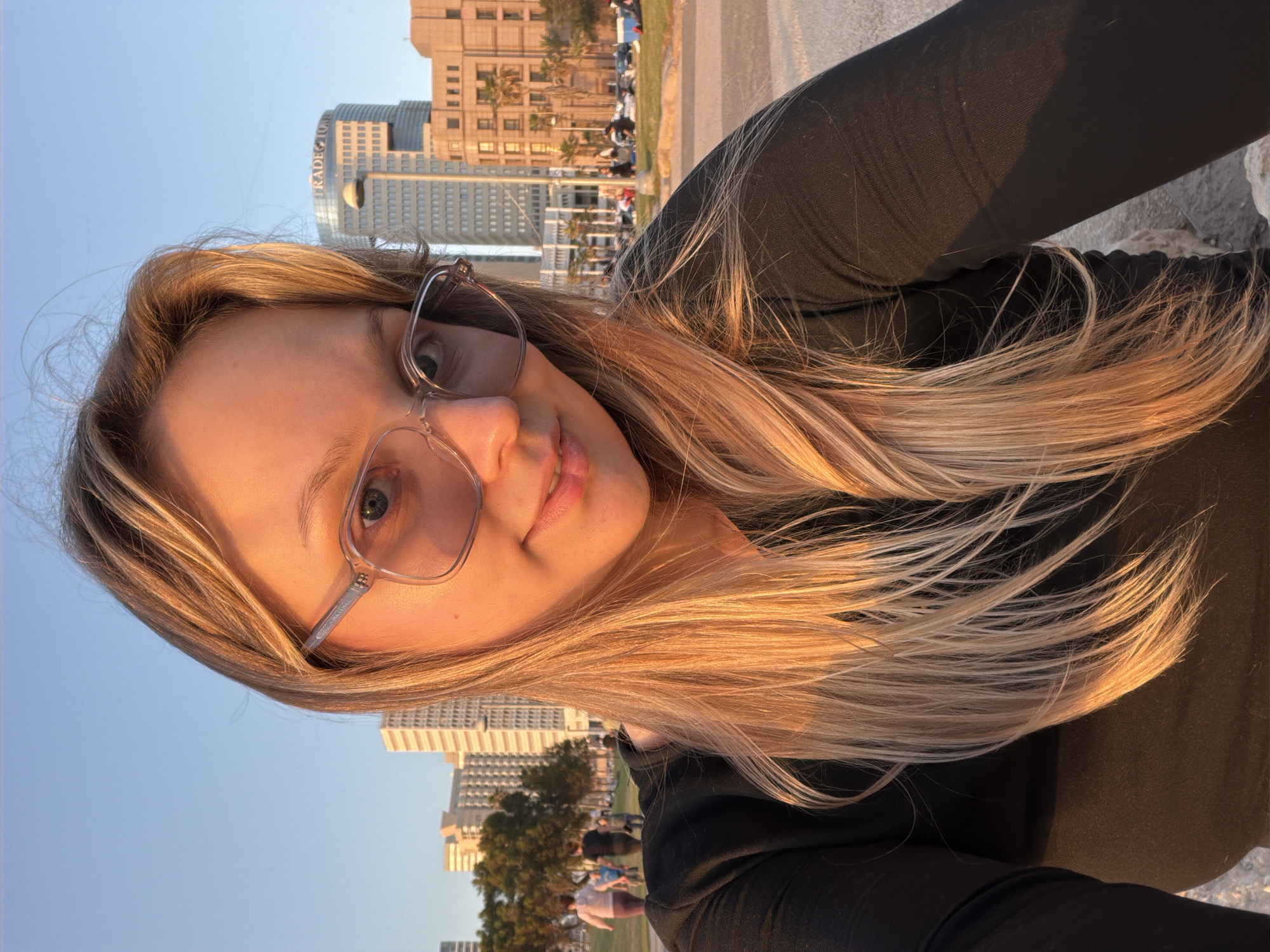 A medium shot depicts a fair-skinned female with blonde hair, wearing glasses, and a black long-sleeved shirt. The background reveals a cityscape, likely Tel Aviv, Israel, given the architectural style of the buildings. People can be seen in the background, suggesting a public space or park. The time of day appears to be late afternoon or early evening based on the warm, golden light.