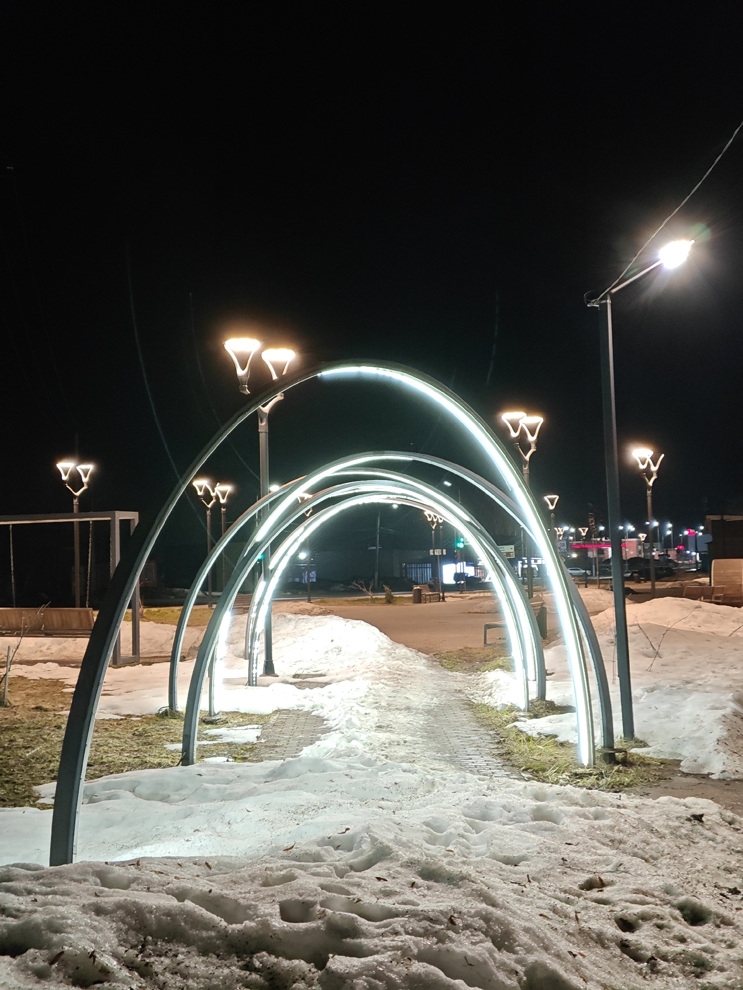 The image depicts an illuminated pedestrian walkway at night in what appears to be a park or public space. The location is likely in Chaikovsky, Russia, based on visual characteristics typical of Russian urban landscapes.
The walkway is defined by a series of arching metal structures, each lined with a light source, creating a tunnel effect. The ground on either side of the walkway is covered in snow, with patches of grass visible where the snow has melted. The walkway itself is paved, with some snow and ice present.
In the background, streetlights illuminate a street with buildings and vehicles. The overall lighting suggests it is nighttime. There are no people clearly visible in the image. Other details visible in the image include standard streetlight poles, a swing set, and benches.