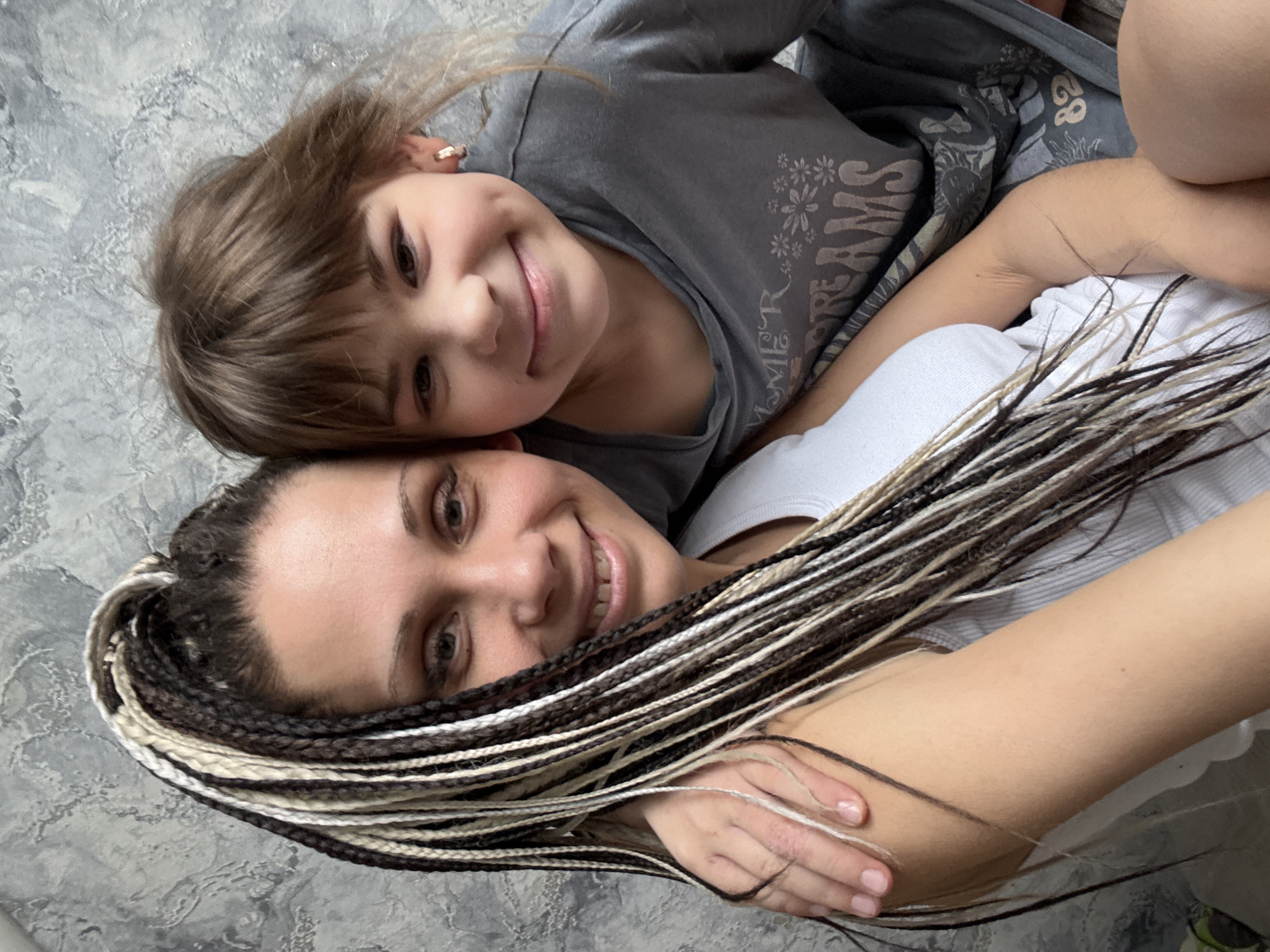 A mother and daughter are lying down together, smiling at the camera. The daughter is in a gray shirt with a floral design, and the mother is in a white shirt. The mother has black and blonde braids. The location is Unknown, Unknown.