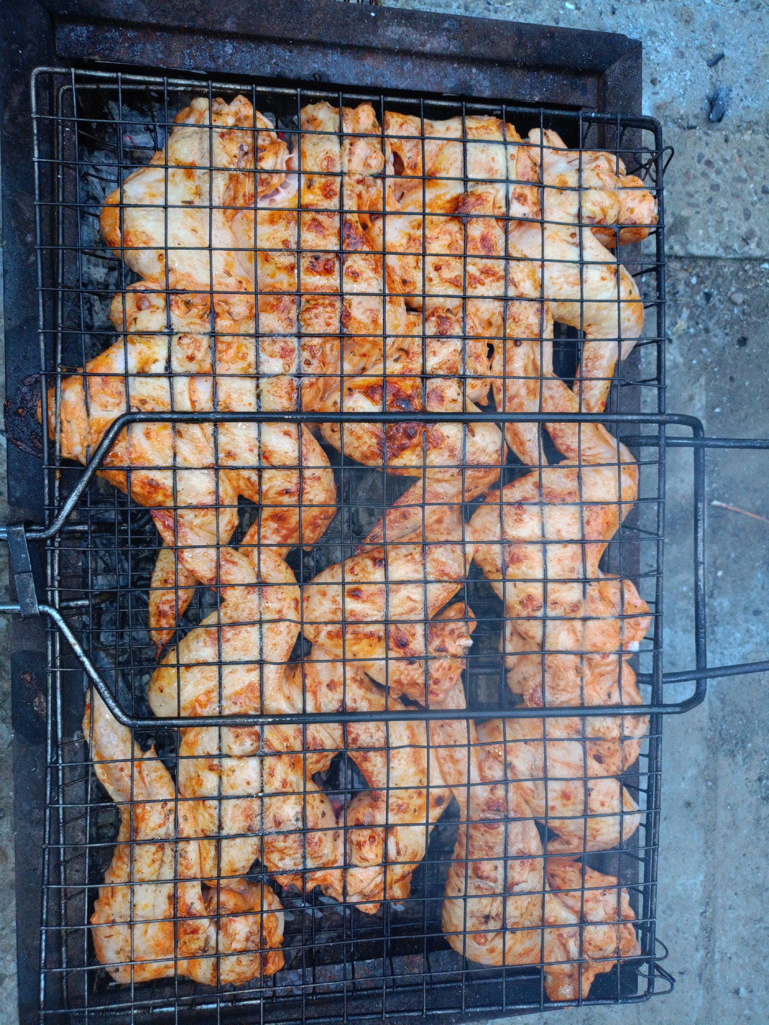 A high-angle, close-up shot shows a metal grill filled with chicken wings being cooked over coals. The chicken wings are seasoned and have a reddish-orange hue from the spices, with some areas showing charring from the grill. The grill is placed on top of a dark metal barbecue grate, which sits above glowing red coals. The background consists of a textured, grey concrete surface, indicating an outdoor setting. The scene appears to be an informal outdoor cooking event in Baurci, Moldova.