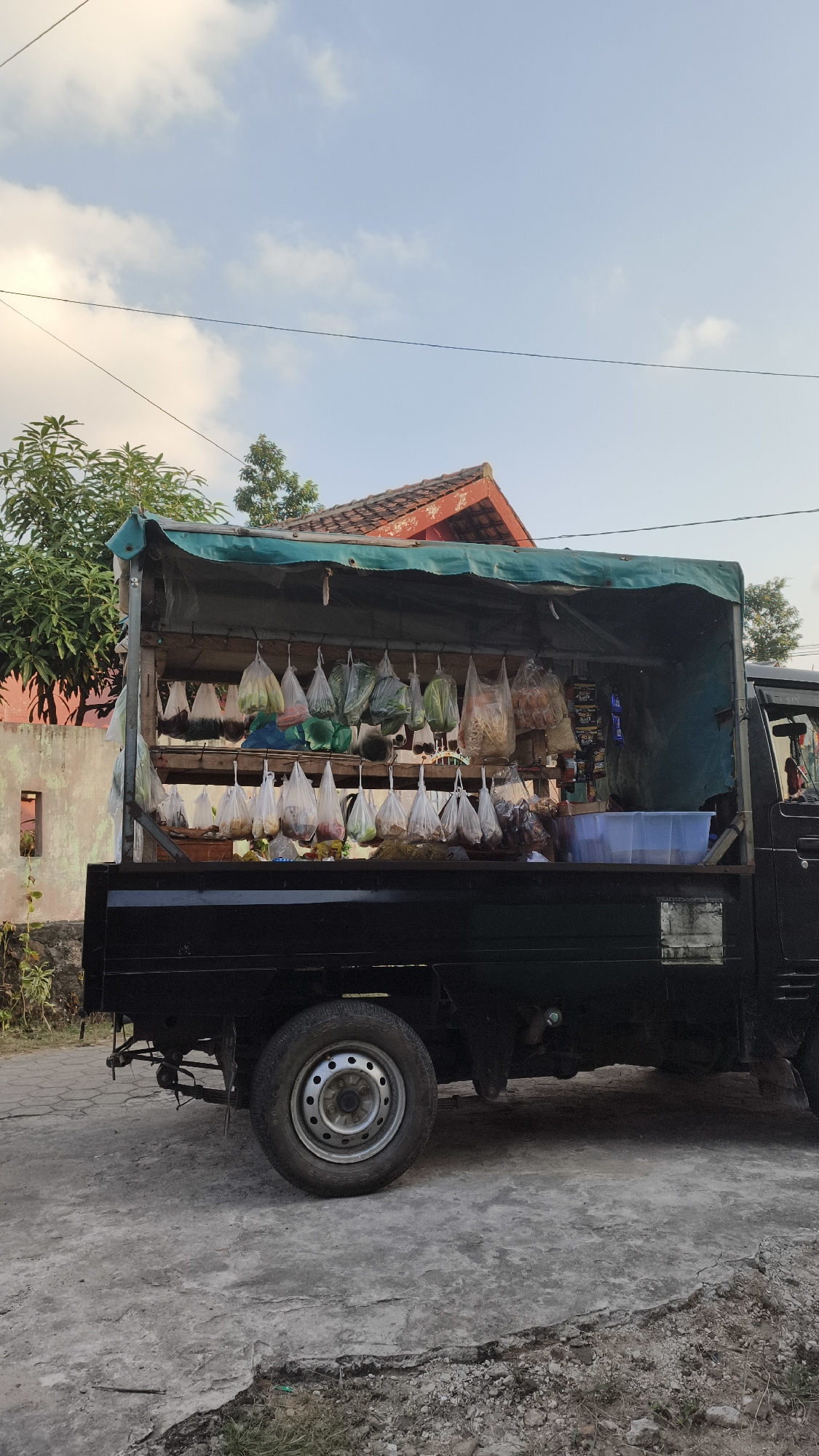 A black pickup truck is parked on a concrete road in what appears to be a residential area. The truck bed is outfitted as a mobile food stall, covered by a green tarp. Numerous plastic bags filled with various food items, such as vegetables and snacks, are hanging from racks inside the truck bed. A blue plastic container is also visible in the truck. Behind the truck, a building with a red tiled roof and an orange wall can be seen, along with some trees and a blue sky with scattered clouds. The scene suggests a typical street market setup in a city like Bandar Lampung, Indonesia, where vendors often use vehicles to sell their goods.