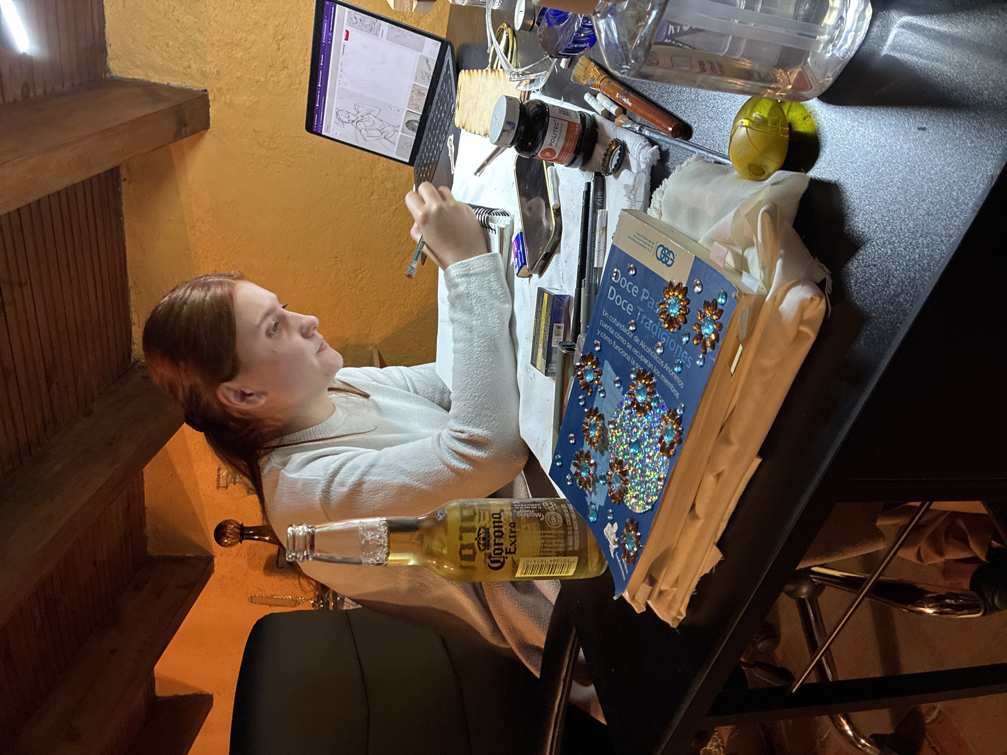 A woman sits at a desk facing a laptop with a drawing on the screen. She is wearing a white sweater and holding a pen. On the desk, there is a beer bottle, a book titled "Doce Pasos Doce Tradiciones," a bottle of ink, brushes, and other art supplies. The location appears to be indoors, possibly in a room with wooden beams and a textured wall. There is no indication of the location being Cañada de Madero, Mexico, within the image.