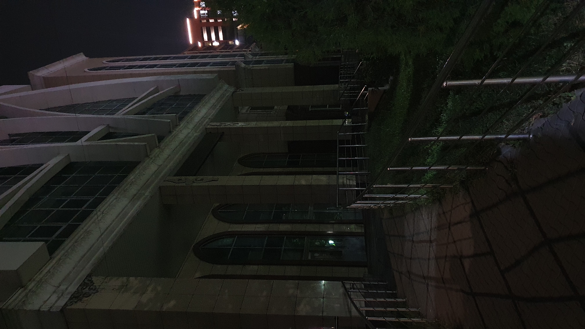 An exterior view of a large, modern building in Almaty, Kazakhstan, is captured at night. The building features a facade of light-colored stone or tile, accented by large, arched windows and vertical columns. A paved walkway, lined with metal railings, leads towards the entrance. The walkway casts long shadows from an unseen light source, creating a dramatic effect on the dark pavement. Adjacent to the walkway, a grassy area is visible, bordered by dense foliage. In the background, another building with illuminated windows and a more contemporary design stands tall against the night sky. The overall scene suggests a quiet, possibly institutional or commercial, setting in the city.