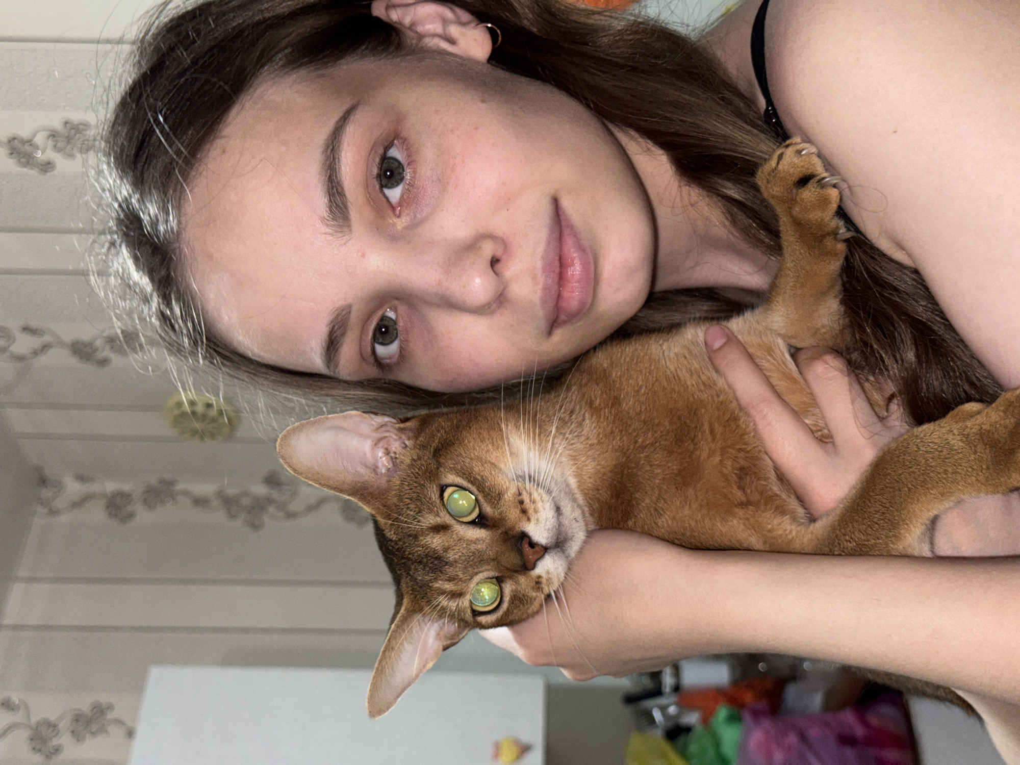 A young woman with brown hair and green eyes, wearing a black strap top, holds a reddish-brown cat with green eyes. The woman is looking directly at the camera, and the cat is nestled in her arms, looking towards the left. The background shows a wall with a floral pattern and what appears to be a kitchen cabinet. The lighting suggests an indoor setting. The image does not provide any information about the location in the city of Chaikovsky, Russia.