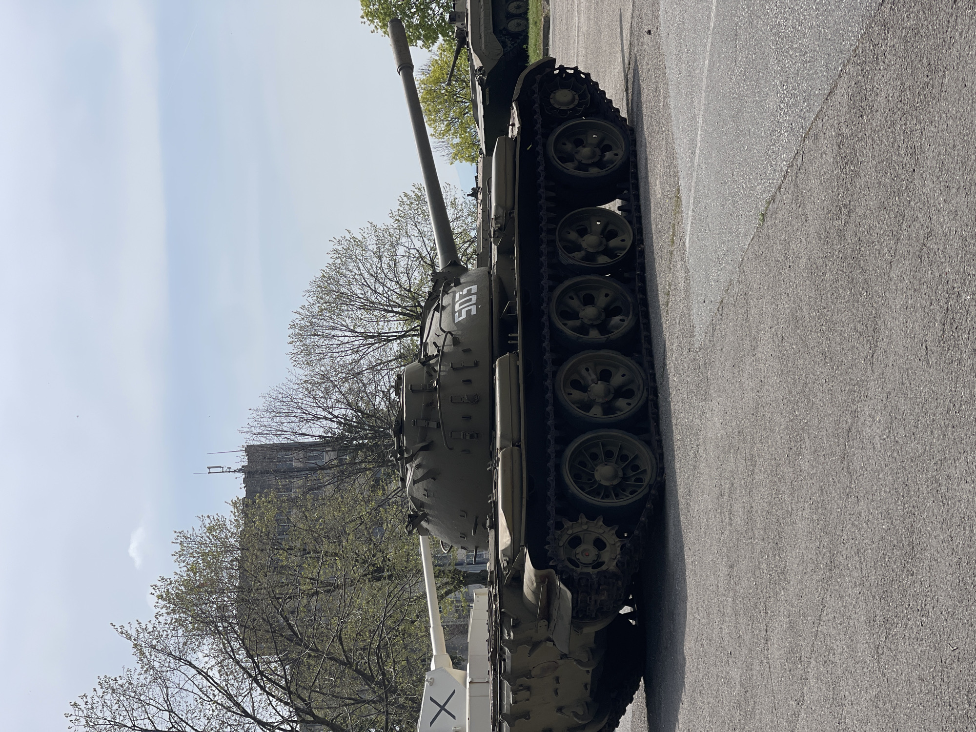 The image shows two tanks on display in Veliko Tarnovo, Bulgaria. The tanks are olive green. The tank in the foreground has the number "503" painted on the turret and a white stripe with a black "X" painted on the front. A building can be seen in the background through the trees. The tanks are parked on an asphalt road. The sky is partly cloudy.