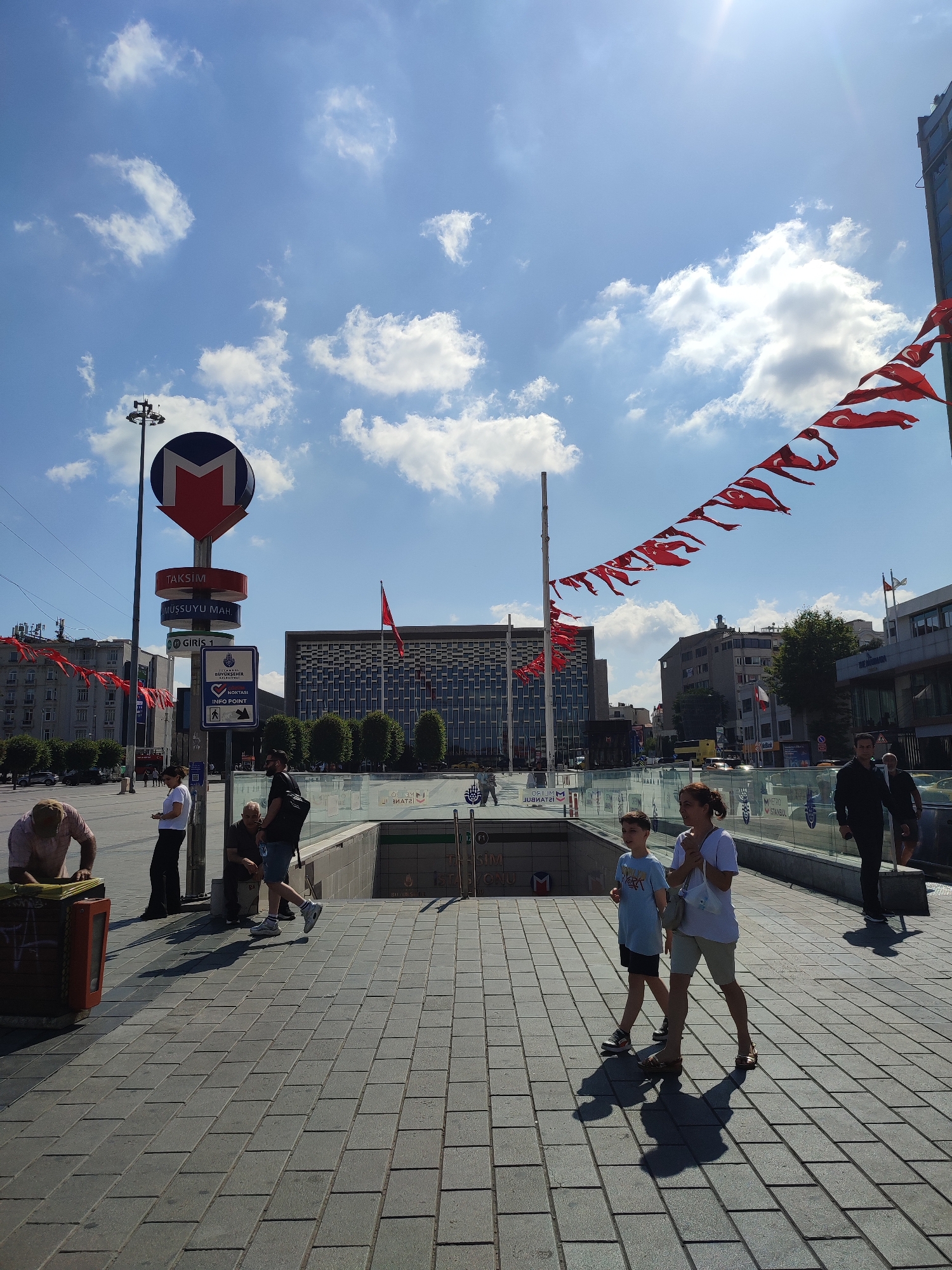 The image depicts a sunny day scene at Taksim Square, Beyoğlu, Turkey. In the foreground, a paved walkway is occupied by several individuals. To the left, a man in a pink shirt bends over a brown trash bin. Further back, a woman in a white t-shirt and black pants stands near a metro sign. A man in a dark t-shirt and shorts, carrying a black backpack, is seated on a low wall, with a water bottle near him. A woman and a young boy, both walking towards the right, are in the mid-ground. The woman wears a white t-shirt and beige shorts, while the boy is in a light blue t-shirt and dark shorts.
Dominating the center background is a large, modern building with a grid-like facade, identified by signage as the Atatürk Cultural Center. To the left of this building, a prominent metro station sign is visible, featuring the letter "M" within a circle and indicating directions for "TAKSIM" and "MÜSSÜYÜ MAHL". An informational sign points to an "INFO POINT".
The sky is blue with scattered white clouds, and the sun is high, casting distinct shadows on the pavement. Red Turkish flags, strung together in a garland, are draped across the upper portion of the frame, extending from the right side towards the center. Other buildings, trees, and traffic are visible in the distance. The overall scene captures a public space with active pedestrian movement and prominent civic landmarks.