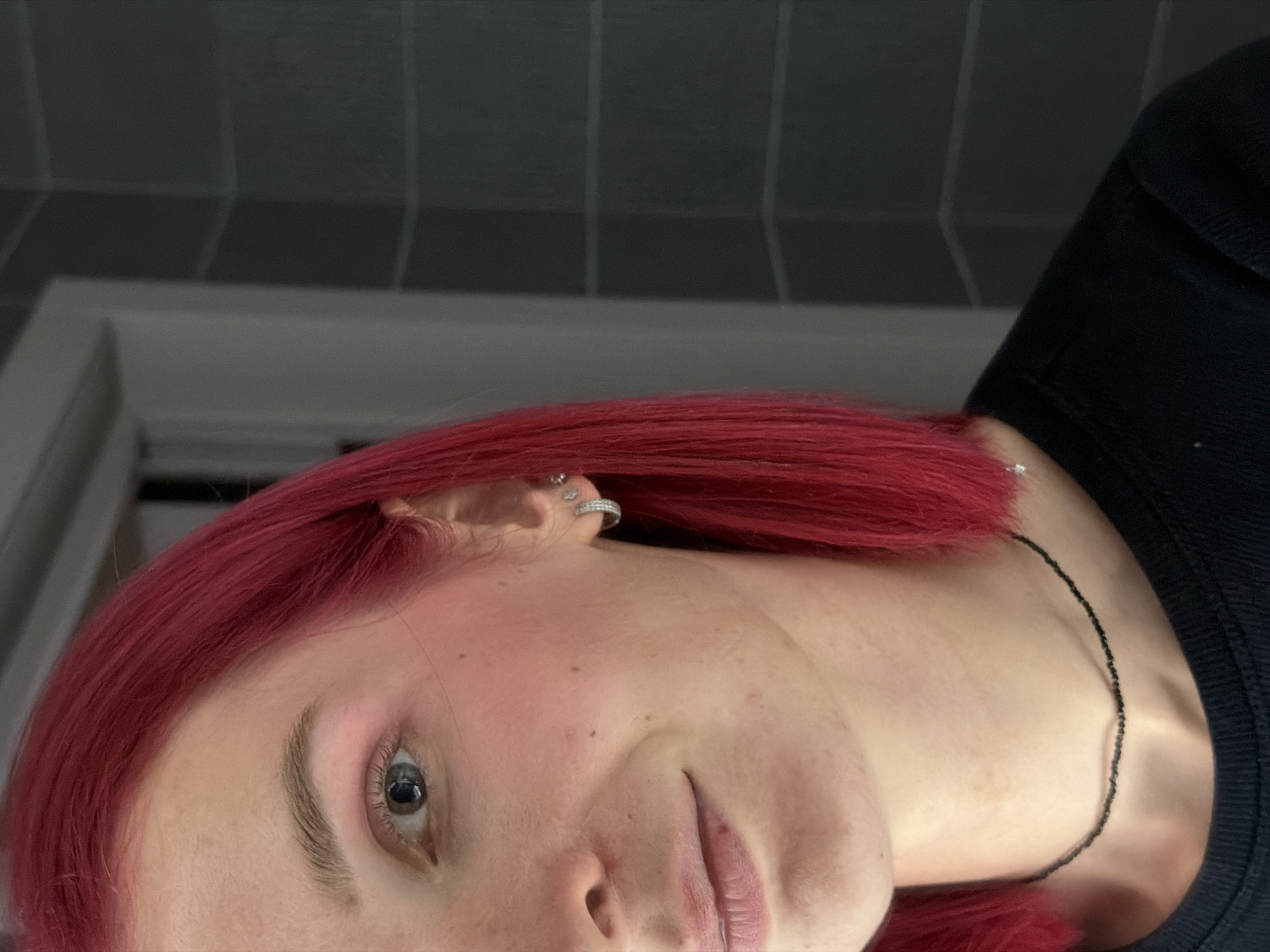 A close-up, tilted portrait-style photograph features a person with bright red hair, positioned indoors. The subject's face occupies a significant portion of the frame, tilted with their head leaning to the left, looking directly towards the camera.
The person has fair skin and visible light eyebrows. One eye is clearly visible, with a light iris color, possibly green or hazel, and subtle eye makeup. Their lips are slightly parted. Several silver-colored piercings are visible on their ear, including a prominent hoop on the helix and another smaller stud. A thin, black beaded necklace is visible around their neck, partially covered by their red hair and a dark-colored garment, likely a shirt or sweater.
The background is simple and somewhat indistinct, suggesting an indoor setting. It features a dark grey tiled or paneled surface at the top, likely a wall or ceiling, with a lighter, blurry structure below it, possibly a doorframe or another architectural element. The lighting is soft and appears to be indoor, without strong shadows or bright natural light cues. The overall mood is neutral, focusing on the individual's appearance. There is no visible text in the image. The location context for the image is Chaikovsky, Russia.
