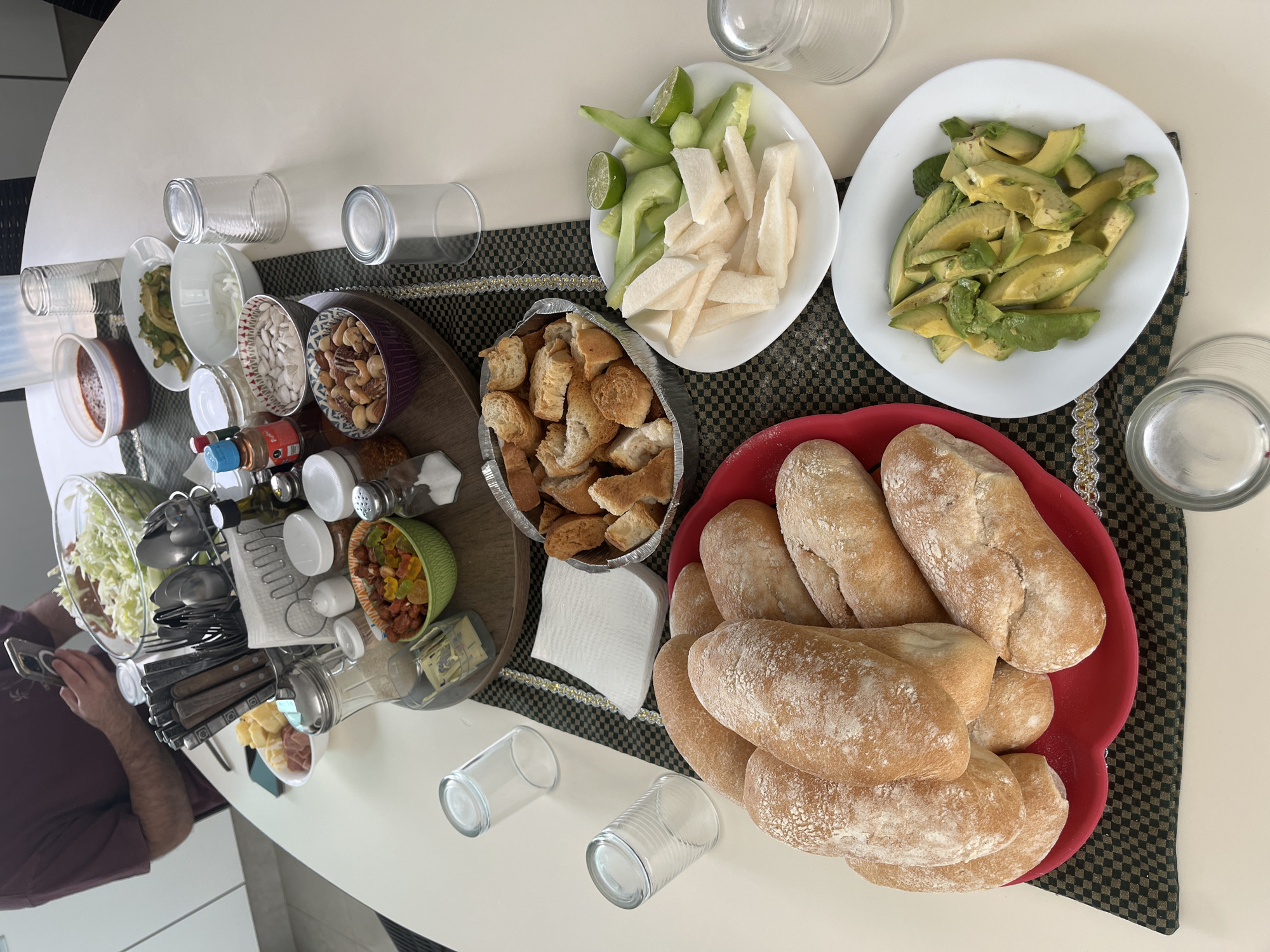 A high-angle shot captures a laden table in San Bernardino Tlaxcalancingo, Mexico. At the table's center, on a black and gold placemat, lie various dishes: a red bowl overflowing with bread rolls, a bowl of toasted bread pieces, a plate of sliced jicama and cucumber with lime wedges, and a plate of sliced avocado. Further down the table, closer to the camera, sits a large bowl of shredded lettuce, various utensils, jars of condiments, and a bowl of what appears to be hard candy or dried fruit. Scattered around the dishes are several empty glass tumblers. A person wearing a maroon shirt is partially visible in the lower left corner of the image, holding a smartphone and likely taking the picture. The table is white, reflecting the overhead light.