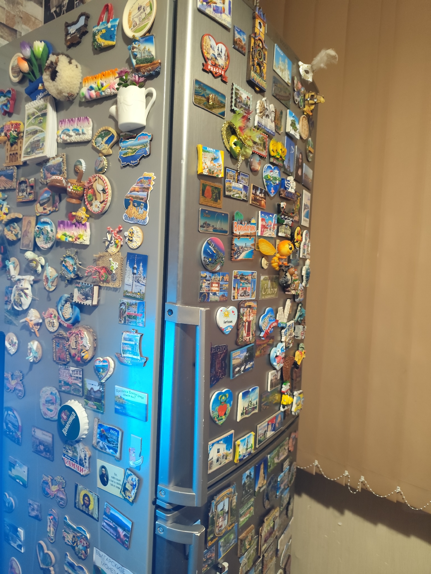 The image shows a refrigerator covered with numerous magnets, most of which appear to be souvenirs from various travel destinations. The refrigerator is silver and positioned against a wall with a beige textured covering. The lighting in the scene is uneven, with a blue light source illuminating the right side of the refrigerator door and the lower portion of the image. Several magnets depict landmarks, cityscapes, and cultural motifs. Some magnets are rectangular, while others are shaped like hearts, bottles, or other objects. The collection suggests a history of travel and memory keeping. No people are visible in the frame. The context of the city of Montana, Bulgaria is not directly indicated by any visual elements in the image.
