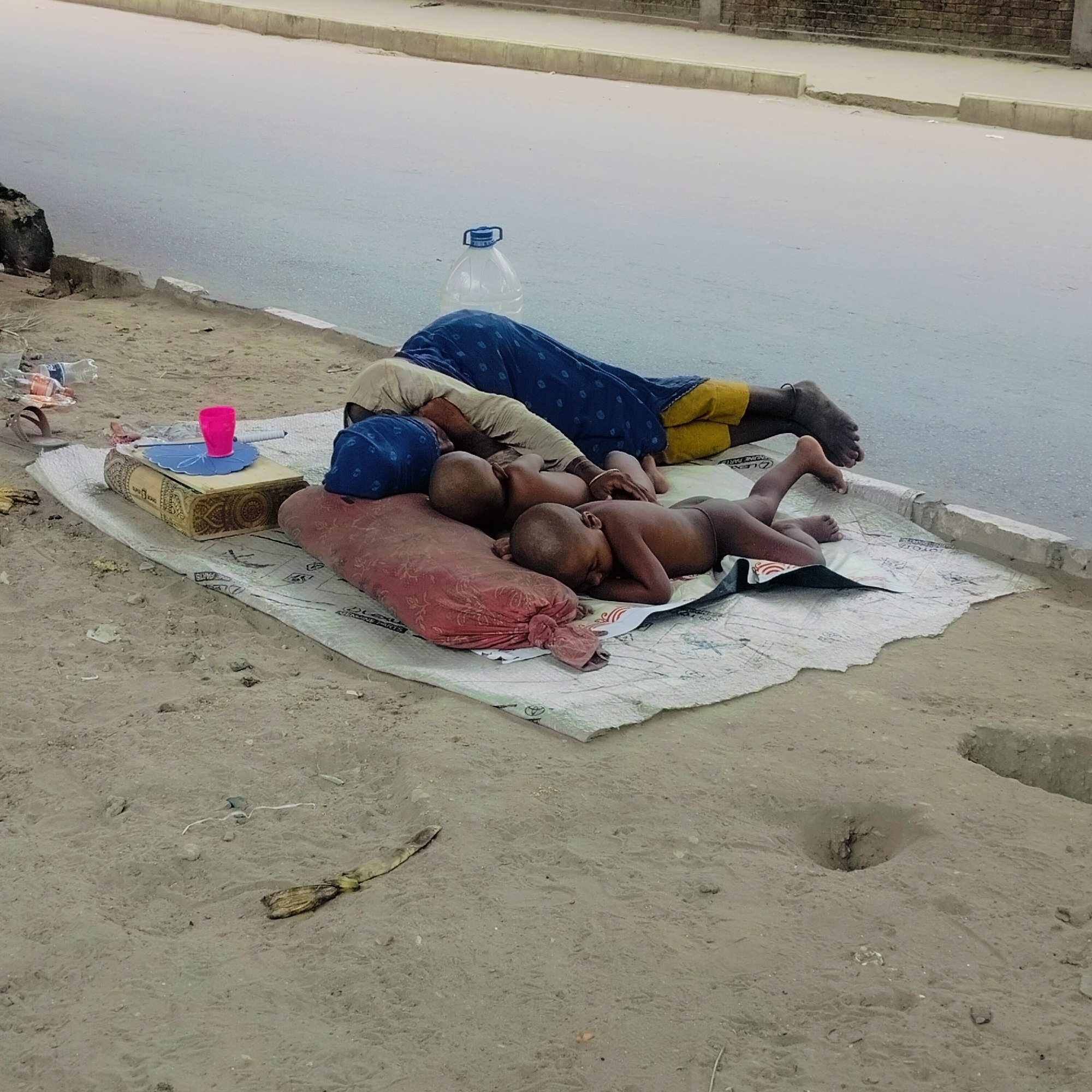 In an urban environment of Unknown, Unknown, two children and an adult lie on a white plastic sheet next to a paved road. The road is grey, and a curb separates it from the sandy ground where the family rests. The adult, dressed in a tan top, blue cloth, and yellow shorts, is lying prone near a large, clear plastic water bottle. Two children, with shaved heads, are lying on the sheet. One rests their head on a red pillow. Next to them is a rectangular, decorated box with a pink cup on top. To the left of the sheet are scattered pieces of trash. A dark object is in the top left corner of the image. In the foreground, the sandy ground is bare except for a stick and a small hole.