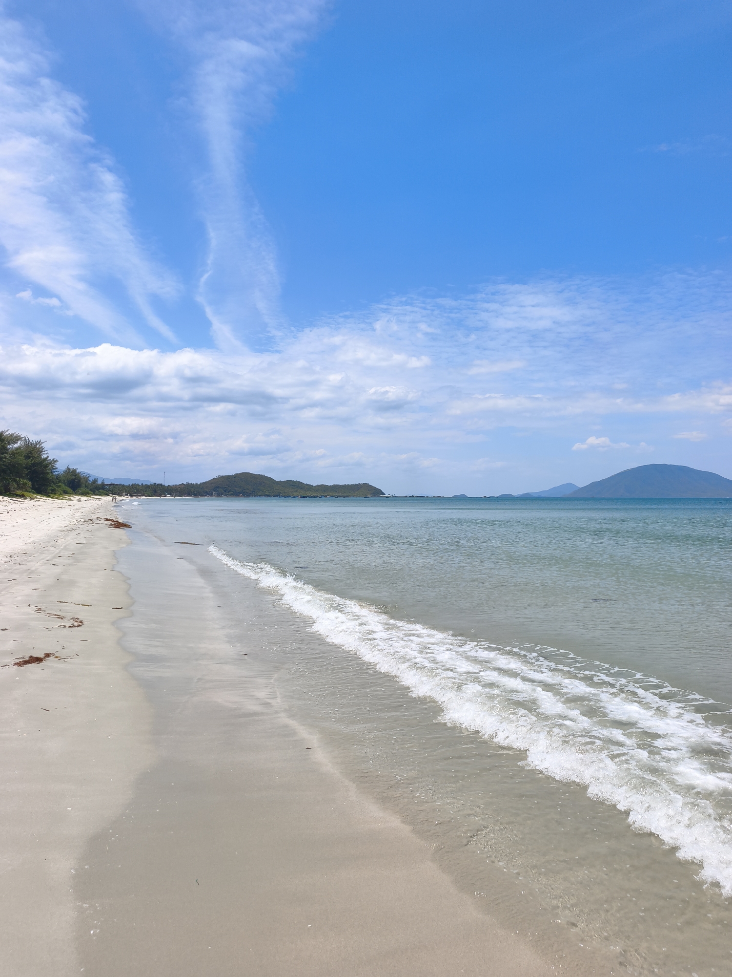 A wide, sandy beach stretches along the coast, meeting a calm, blue sea. Gentle waves with white foam lap at the shore. In the distance, a line of lush green hills and mountains borders the horizon. The sky is a bright blue, dotted with wispy white clouds. A few scattered pieces of dark seaweed lie on the wet sand near the water's edge. The scene is serene and evokes a sense of natural beauty. The location is identified as Dong Ninh Hoa, Vietnam.