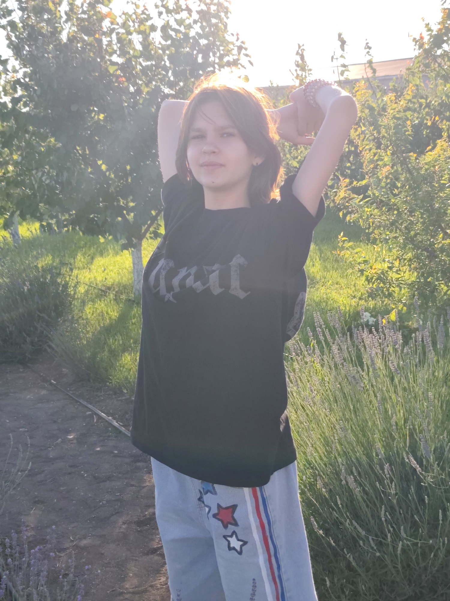 A young person with medium-length brown hair stands in an outdoor setting. They are wearing a black t-shirt with a gothic-style text graphic and light-colored pants adorned with red and white stars and stripes. Their arms are raised above their head, with hands clasped behind their neck.
The scene appears to be a garden or yard, with trees and lush green foliage visible in the background. A patch of lavender is in bloom to the right of the subject. The lighting suggests it is late afternoon or early evening, with the sun casting a warm glow and creating some lens flare. The ground in front of the subject is a dirt path. The overall mood seems relaxed and peaceful. No specific event is discernable, but the posture might indicate stretching or posing. The text on the t-shirt is in a stylized font and appears to be a word or name in a gothic script, but it is not clearly legible.