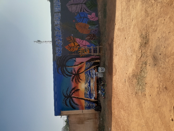 This image depicts an outdoor daytime scene in Kirana, Nigeria, centered on a long, concrete wall serving as a mural canvas. The ground in the foreground and midground consists of reddish-brown dirt with sparse, dry vegetation.
The wall, approximately 2 meters in height, is divided into two distinct mural sections. The left section portrays a tropical sunset scene with two prominent black silhouettes of palm trees flanking a sun setting or rising over a body of water with white-capped waves. The sky transitions from orange and yellow hues near the sun to a deep blue at the top. Below this mural, on the dirt ground, a black plastic bag, several small pieces of refuse, and two white plastic paint buckets are visible.
The right section of the wall features a dark grey background with stylized text in blue and white, reading "WELCOME 2 BLUE VALLE". Below the text, several large, stylized leaves are painted in shades of orange, brown, purple, pink, blue, and light green. A small, three-step wooden ladder leans against the wall beneath this mural.
In the far background, against a clear, pale blue sky, the upper structure of a metallic lattice communication tower is visible above the wall. To the far left, behind the wall, a light-colored building structure topped with barbed wire is partially visible. No individuals are present in the frame.