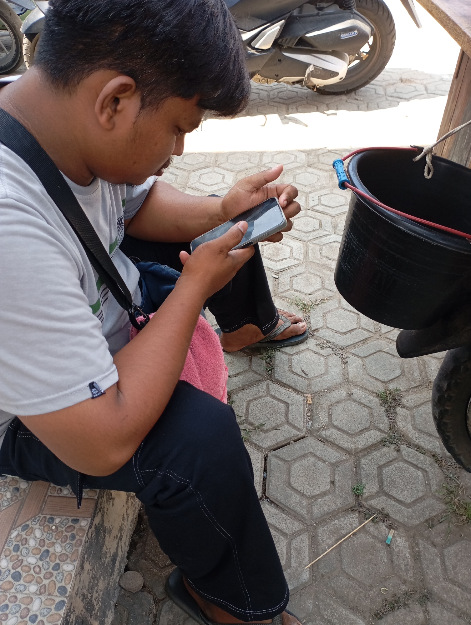 A person is seated outdoors, looking down at a smartphone. The person is wearing a light-colored t-shirt, dark pants, and flip-flops. A pink towel is placed on their lap, and a black strap from a bag crosses their chest. The ground is paved with hexagonal grey tiles. In the background, a portion of a motorcycle and a black bucket with red and blue accents are visible. The scene appears to be in a public space or a casual outdoor setting within Kecamatan Haurgeulis, Indonesia.