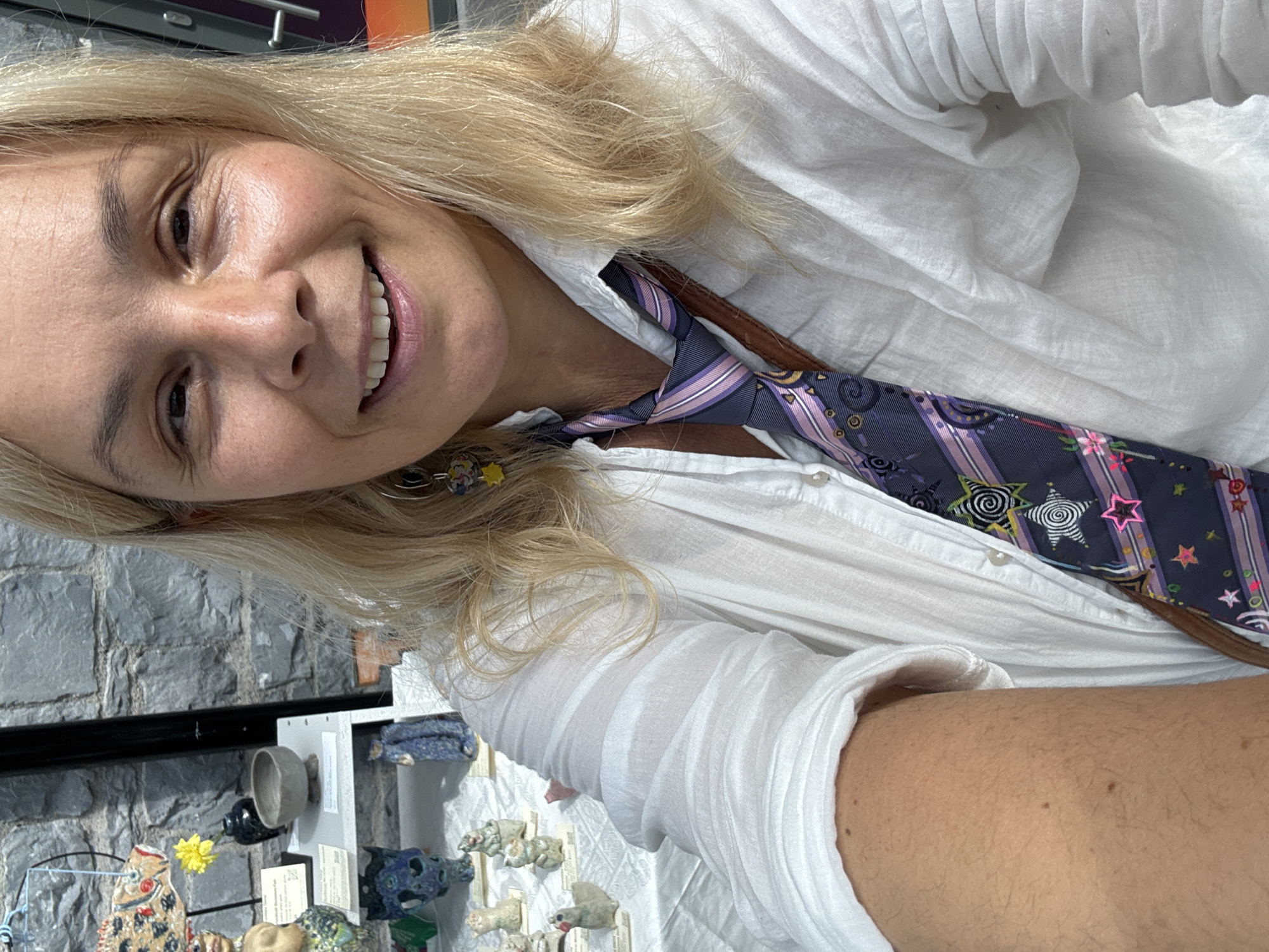 A woman with blonde hair and fair skin is captured in a selfie, smiling at the camera. She is wearing a white collared shirt, unbuttoned at the top, revealing a brown strap over her shoulder and a dark tie with a colorful star pattern. Her left arm, bare and tanned, is positioned in the foreground, suggesting she is holding the camera. Behind her, a display of ceramic art is visible on white tables, with a stone wall in the background. The scene appears to be an outdoor market or craft fair. The setting is likely Kilkenny, Ireland, suggested by the presence of a sign that reads "Kilkenny Design Centre".