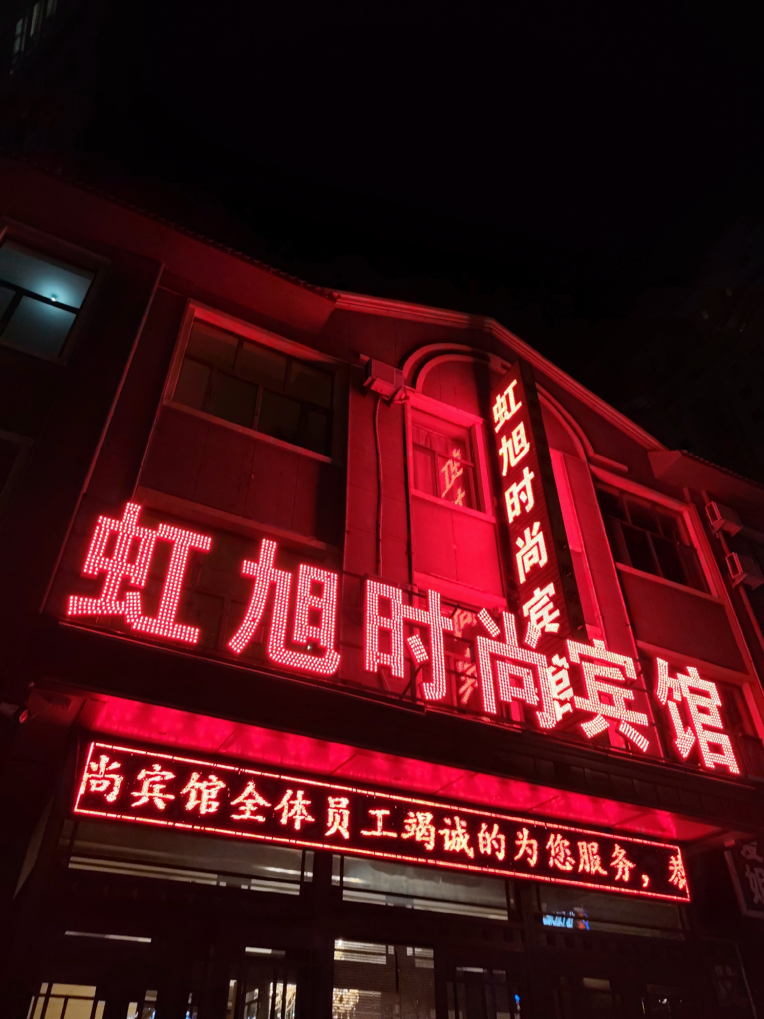 The image captures a nighttime exterior of a building with prominent red neon signage. The building appears to be a commercial establishment, possibly a hotel or a similar service business, given the text on the signs. The largest sign, written in bright red LED characters, reads "虹旭时尚宾馆" which translates to "Hongxu Fashion Hotel." A vertical sign on the right side of the building also displays "虹旭时尚宾馆" vertically. Below the main sign, a smaller LED display reads "宾馆全体员工竭诚的为您服务," meaning "All hotel staff are dedicated to serving you." The building has multiple windows, some lit from within with a cool blue light, contrasting with the red exterior lighting. The scene is set against a dark night sky. This establishment is located in Lindian, China.