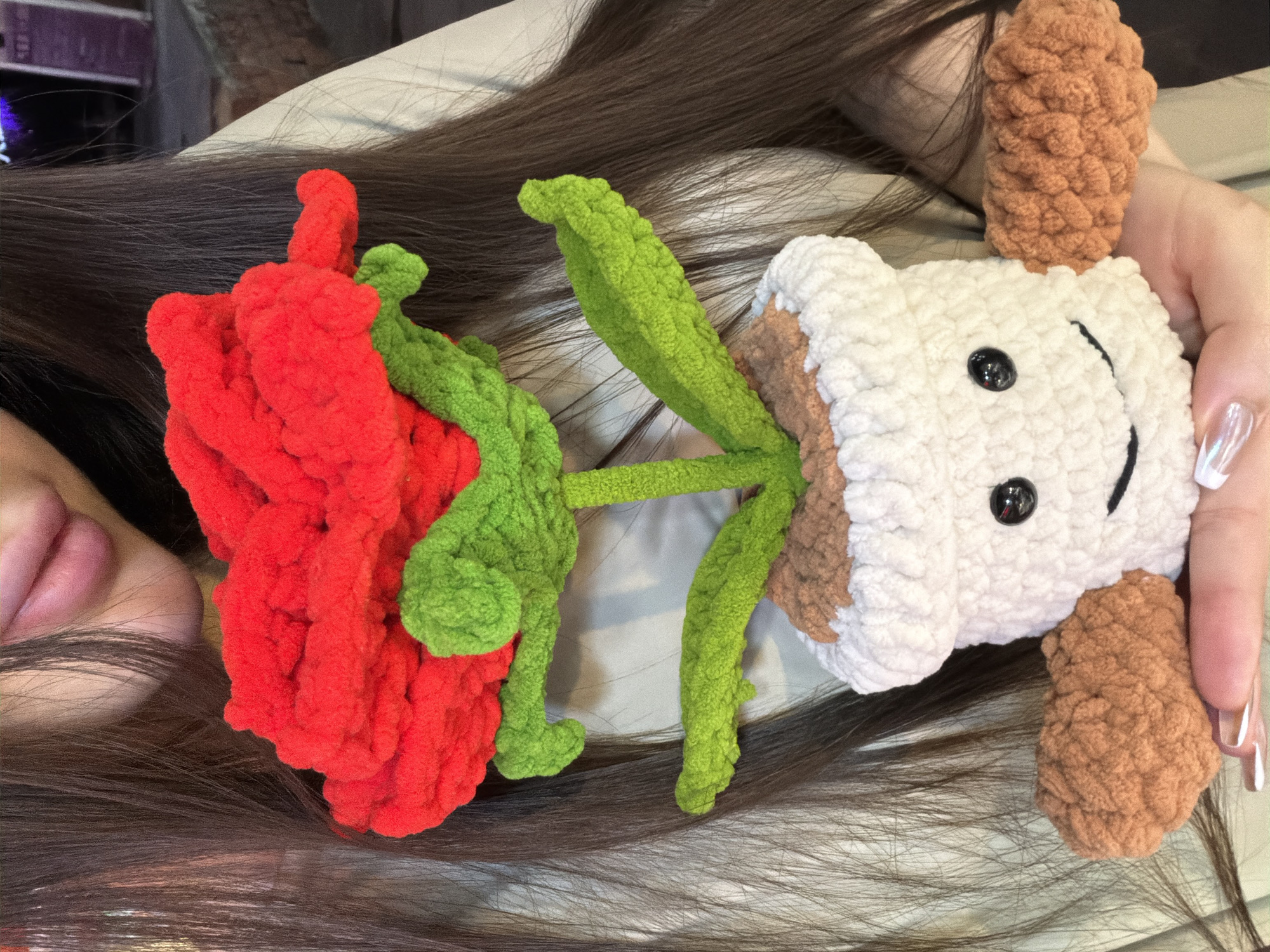 A person is lying on a bed with a crocheted dog and rose toy on their head. The rose is red with green leaves, and the dog is white with brown ears. The person has long brown hair and is wearing white nail polish. The location is likely indoors in Chongqing High-tech Zone, China.