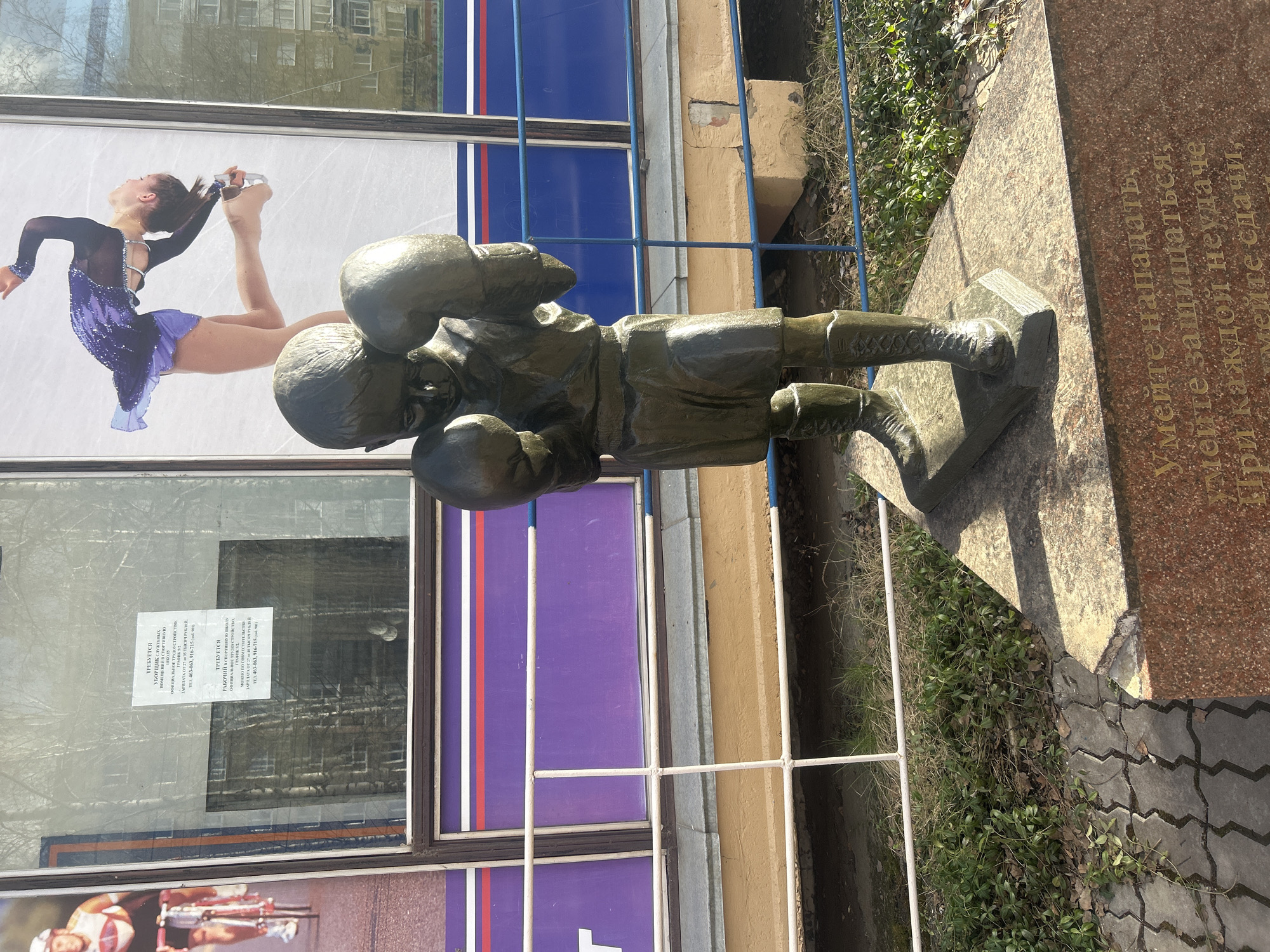 Here's a forensic description of the image:
**Scene:** Outdoor location in what appears to be Izhevsk, Russia.
**Objects and Subjects:**
* Bronze statue of a young boxer in a fighting stance, positioned on a stone base/plinth.
* Behind the statue, there is a building facade. The building has large windows displaying advertisements. One visible advertisement features a female figure skater. There is a notice in Russian on the window as well.
* A light metal fence separates the statue from a narrow strip of vegetation.
* The stone base appears to have a plaque with text in Russian.
**Environment:**
* The weather seems to be sunny with the presence of shadows.
* Paved ground is visible.
**Location:** Izhevsk, Russia (based on the Cyrillic script on the window and stone base).