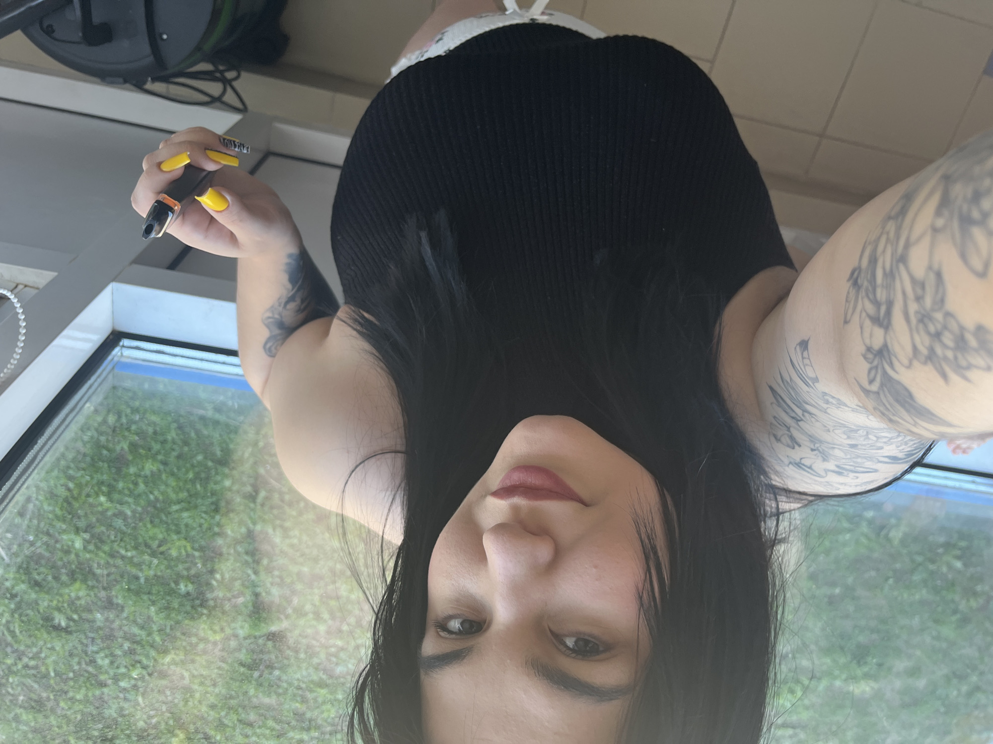 A selfie shot angled downward at a woman with black hair wearing a black tank top, partially cropped at the top, with white trim suggesting underwear or shorts. The woman is holding a vaping device with yellow nail polish visible on her fingers. The subject has tattoos on both arms. The background shows a window with an exterior view of greenery suggesting a yard or park. No information can be determined to confirm the city location.