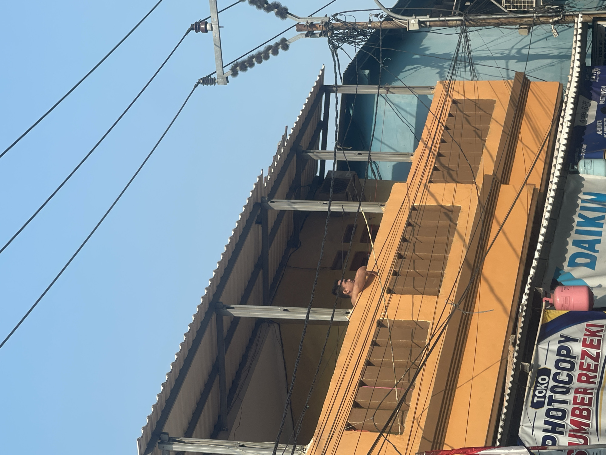 The image depicts an outdoor scene in Bekasi, Indonesia. A man is leaning over an orange balcony, looking to his left. Behind him, a building with a corrugated metal roof is visible. Electrical wires crisscross the sky, with a utility pole on the right side of the frame. Below the balcony, a banner for "TOKO PHOTOCOPY SUMBER REZEKI" is displayed, along with a pink gas cylinder. Another building, painted blue, is visible in the background. The sky is clear and blue.