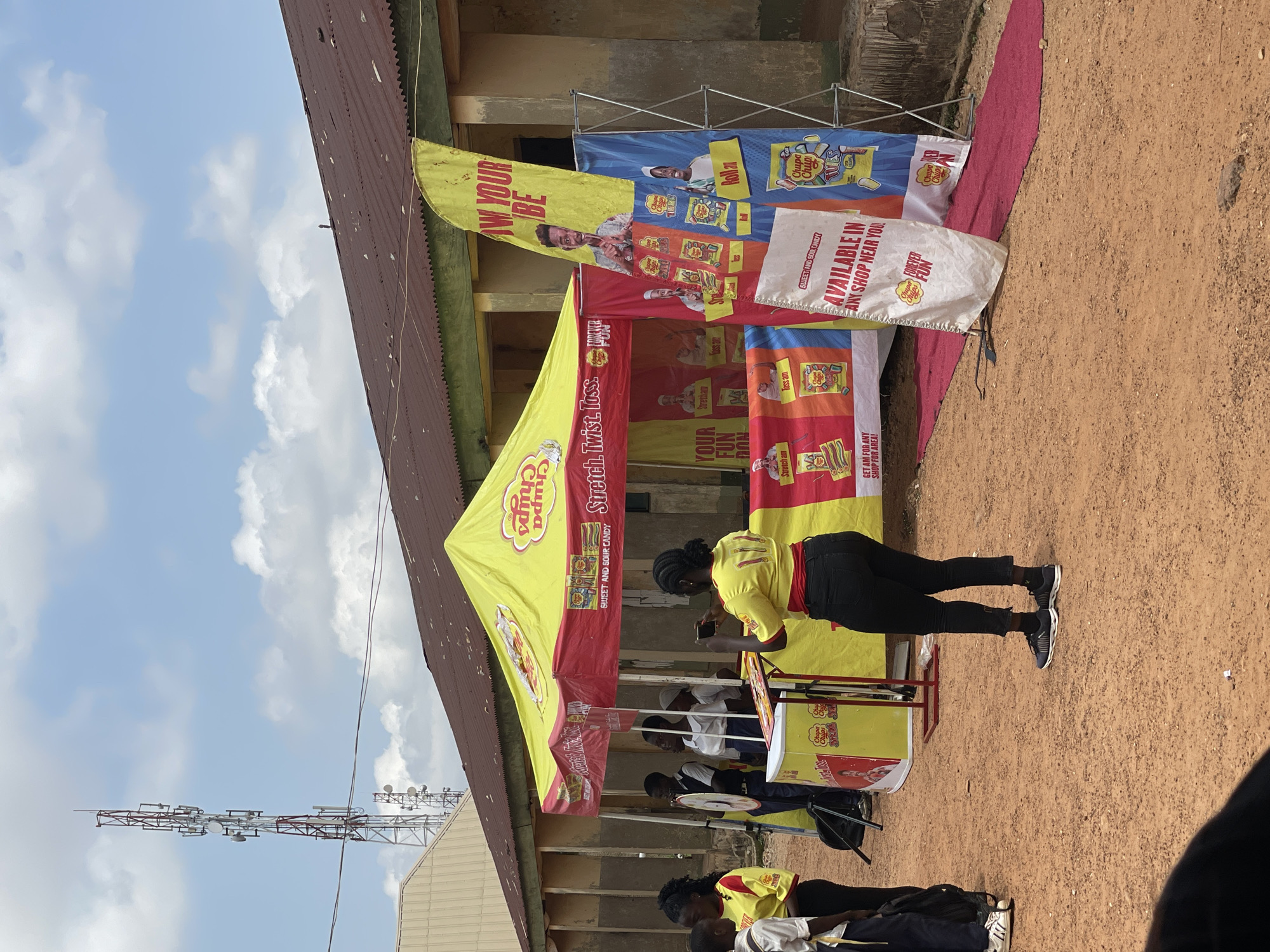 This is a vertical, eye-level, outdoor shot of a promotional event taking place in a dusty, open area, possibly a market or community space. The time of day appears to be midday, with bright sunlight casting shadows. A large, yellow and red tent serves as the focal point, adorned with vibrant banners advertising "Chupa Chups" candy. The banners feature images of candy packs and people engaged in playful activities, with text in various colors, including English slogans like "Stretch. Twist. Toss.", "Your Fun.", "Roll on", "Toss on", and "Available in any shop near you!".
Several people are visible, mostly young adults, dressed casually. One woman, wearing a yellow Chupa Chups branded shirt and black pants, is actively engaged, holding a phone as if taking a picture or video. Others, some in similar branded shirts, are positioned behind a counter or table, suggesting they are part of the promotion. The overall atmosphere seems energetic and promotional, with a focus on engaging the public.
In the background, a building with a corrugated iron roof and an older facade is visible. Beyond that, a telecommunications tower stands tall against a partly cloudy sky. The ground is a reddish-brown dirt, typical of arid or semi-arid regions. The presence of the television tower hints at a connection to broadcasting or media events, and the mention of Nigeria in the location context further grounds the scene. The scene conveys a sense of local community engagement with a global brand.