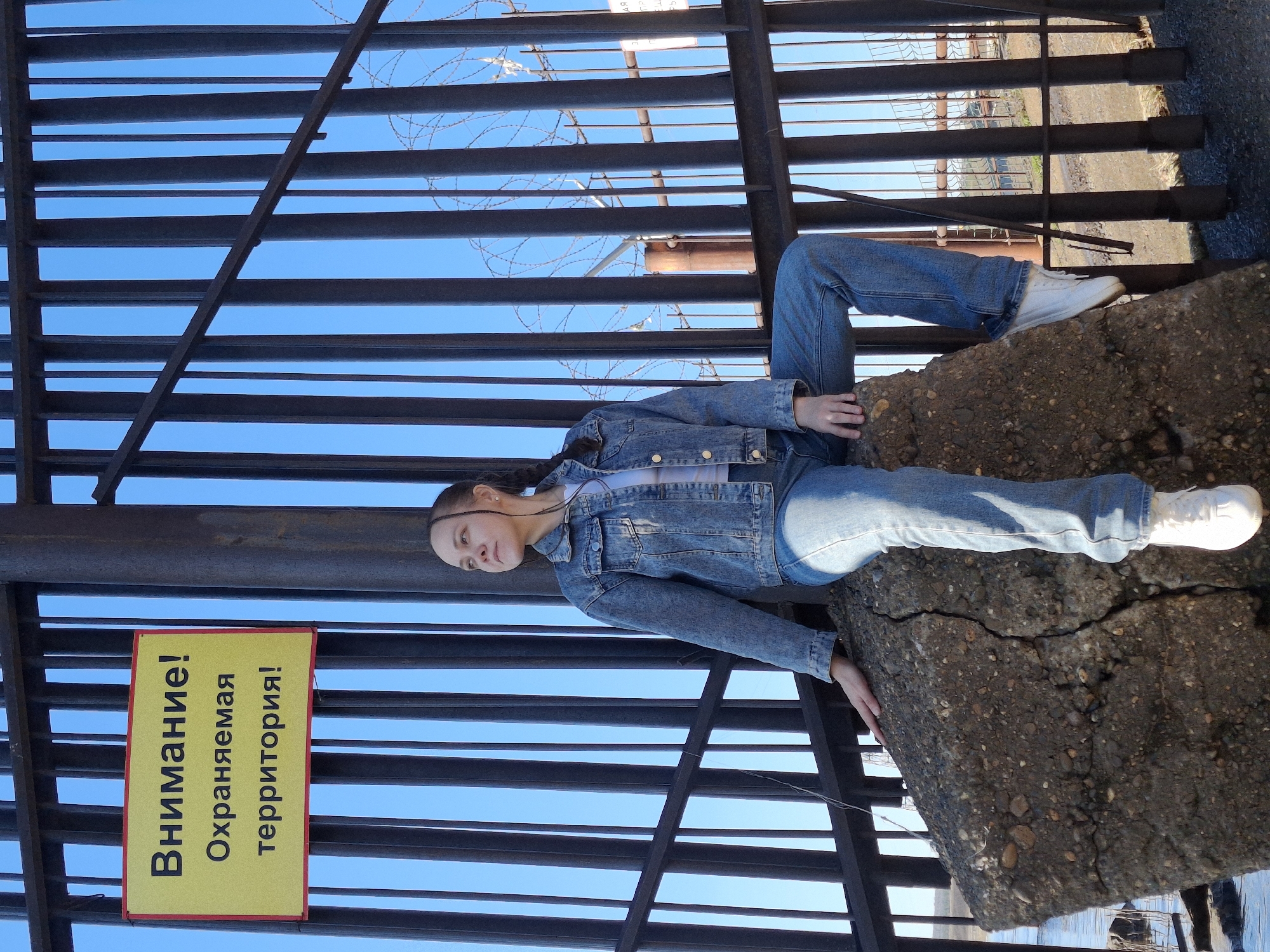 The image shows a young person posing outdoors in Chaikovsky, Russia, on a bright, clear day. The individual, dressed in a light-wash denim jacket, blue jeans, and white sneakers, is lying on their back on a large, weathered concrete block. Their legs are bent, with one foot resting on the block, and their hands are casually placed on the concrete beside them. Their dark hair is styled in braids, and their expression is neutral, looking slightly upwards.
Behind and above the person is a prominent dark metal fence or grille, composed of multiple horizontal slats. A clear blue sky is visible through the gaps in this structure. Barbed wire is visible along the upper section of the fence, indicating a secured area.
Attached to the fence is a yellow rectangular sign with red borders. The text on the sign is in Cyrillic and reads:
"Внимание!" (Vnimanie!) – "Attention!"
"Охраняемая" (Ohranyaemaya) – "Guarded"
"территория!" (territoriya!) – "territory!"
This translates to "Attention! Guarded territory!" The sign, barbed wire, and the industrial appearance of the concrete block and fence all suggest the location is a restricted or protected zone. The time of day is daytime, with bright sunlight and a clear sky.