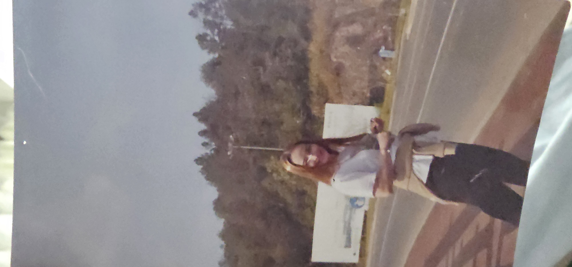 In a medium shot, a young woman with long, light brown hair stands outdoors, smiling slightly at the camera. She is wearing a white t-shirt and dark pants, with a light brown sweater tied around her waist. The woman is standing on a paved surface, possibly a sidewalk or path, with a road visible to her right. Behind her, a large white billboard with some indistinct text and images is visible. Further in the background, a hillside covered in trees and foliage can be seen, with a street lamp rising above the treeline. The sky is a pale blue with some light clouds. The scene suggests an outdoor location, possibly a roadside area, with natural elements and man-made structures. The image is static, capturing a single moment in time. The specific location in Шыңғырлау ауылдық округі, Kazakhstan, is not explicitly depicted, but the presence of a billboard and road implies a developed area.