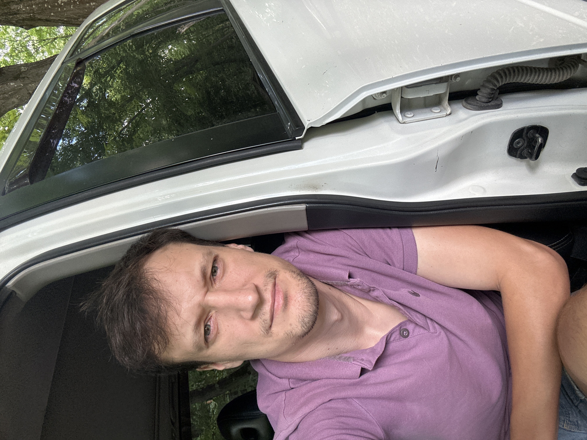 The image is a close-up selfie of a man sitting in the open driver's side door of a white vehicle. The man has brown hair and is wearing a purple polo shirt. His arms are crossed and he is looking towards the camera with a slightly furrowed brow. The interior of the car is visible behind him, as is the exterior of the car door and frame. Through the tinted windows of the car, trees and greenery are visible, suggesting an outdoor, possibly wooded, location. The photo appears to have been taken during the daytime, with natural light illuminating the scene. There is no indication of the specific location within Shatava, Ukraine.