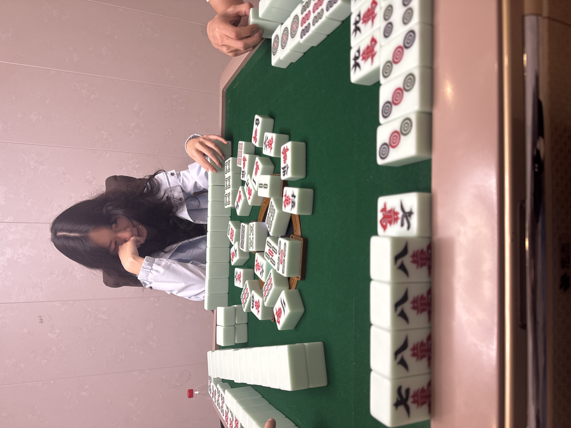 A young woman with long dark hair is resting her head on her hand, looking at a Mahjong game in progress on a green-topped table. The table is covered in Mahjong tiles, arranged in walls and scattered across the playing surface. Another person's hand is visible above the tiles, indicating multiple players. A water bottle with a red cap rests near the bottom left of the table. The table has a glossy pink surface and what appears to be a wooden frame. The background features a pale pink wall with a subtle pattern. The location is likely indoors, based on the lighting and environment. The city of Nan'an District, China, cannot be confirmed.