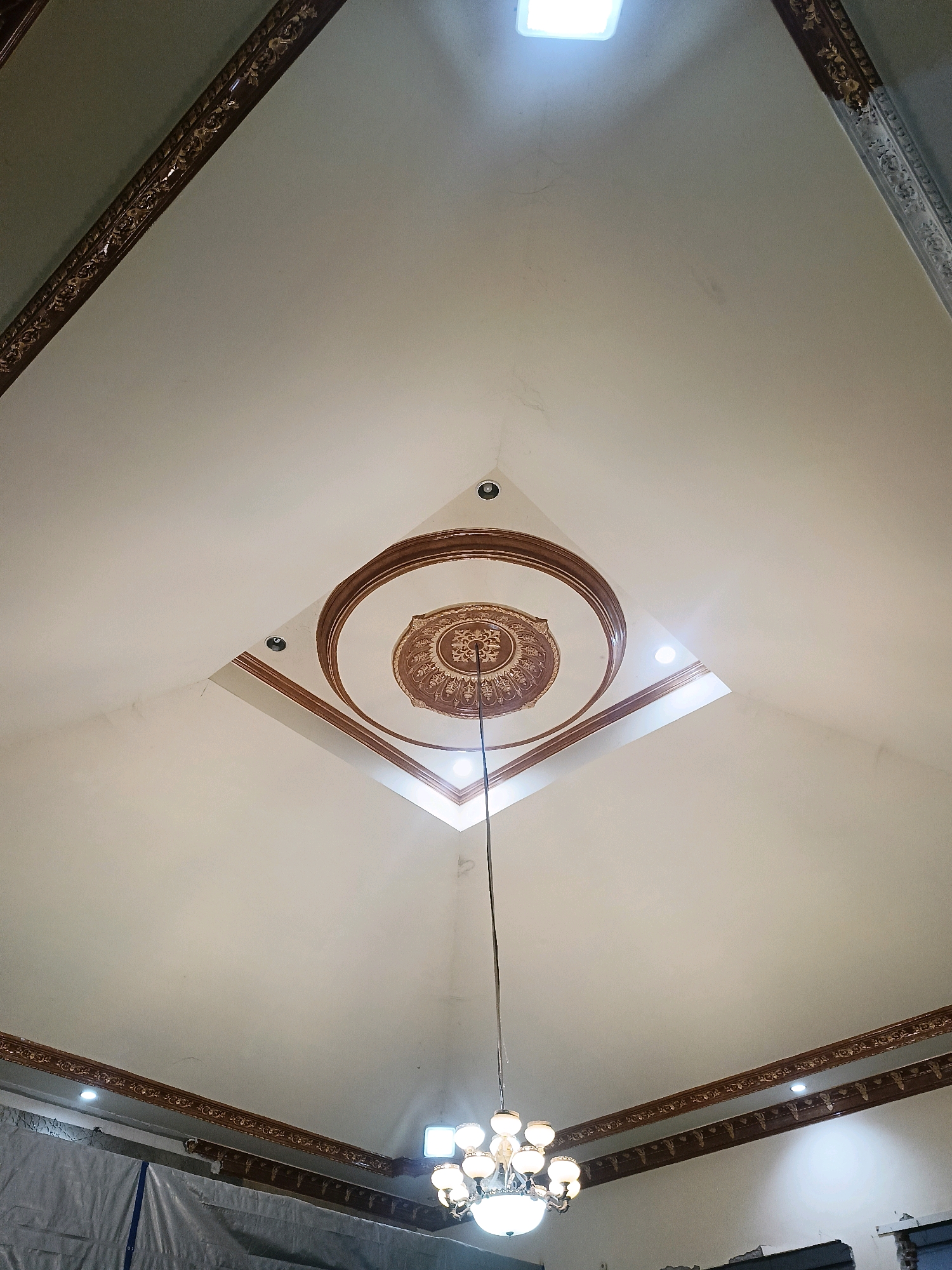 The image shows a ceiling with intricate decorative molding and a central medallion. There is a chandelier hanging from the ceiling and a skylight at the top. The overall impression is one of a well-decorated interior space. The location is identified as Kediri City, Indonesia. The scene is static and depicts an interior architectural detail. There are no people or animals visible. The environment appears to be a building, possibly a home or a public space. The lighting suggests it is daytime, with natural light from the skylight and artificial light from the chandelier and other ceiling fixtures. The image focuses on the ceiling and its decorative elements.