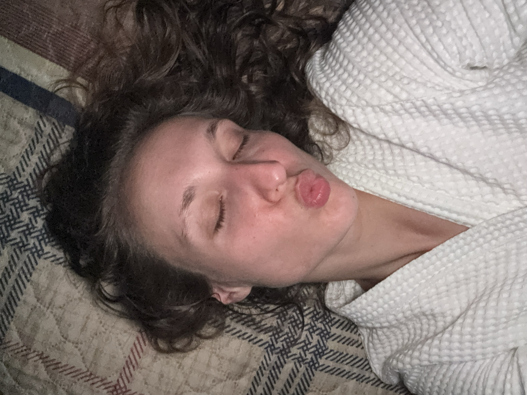 A close-up, overhead shot captures a young woman lying down, her eyes closed and lips pursed in a kiss. She is wearing a white waffle-weave bathrobe. Her dark, curly hair is spread out around her head, and she is lying on a patterned blanket. The scene is likely indoors, possibly in a bedroom. There are no contextual elements within the image that would allow for the identification of the city of Kuyeda, Russia.