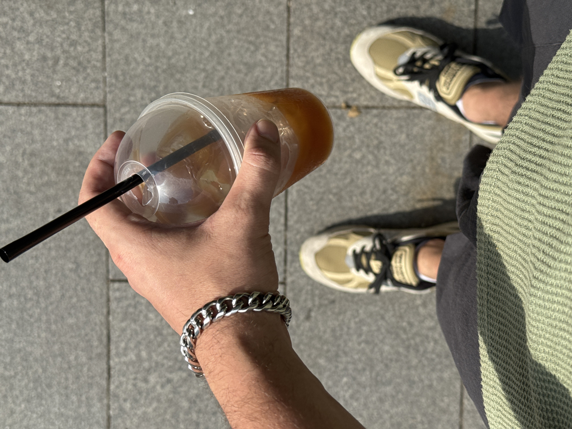 The image captures a first-person perspective of a person walking on a grey paved surface, likely a sidewalk or public walkway. The person is holding a clear plastic cup filled with an iced beverage and a black straw. A chunky silver bracelet adorns the person's wrist. The person is wearing a pair of beige and white sneakers with black laces. Part of dark grey shorts and a light green textured garment, possibly a sweater or jacket, are visible. The environment suggests an outdoor setting in a city, with the paved ground and natural lighting. There is no specific visual information to pinpoint the location to Moscow, Russia.
