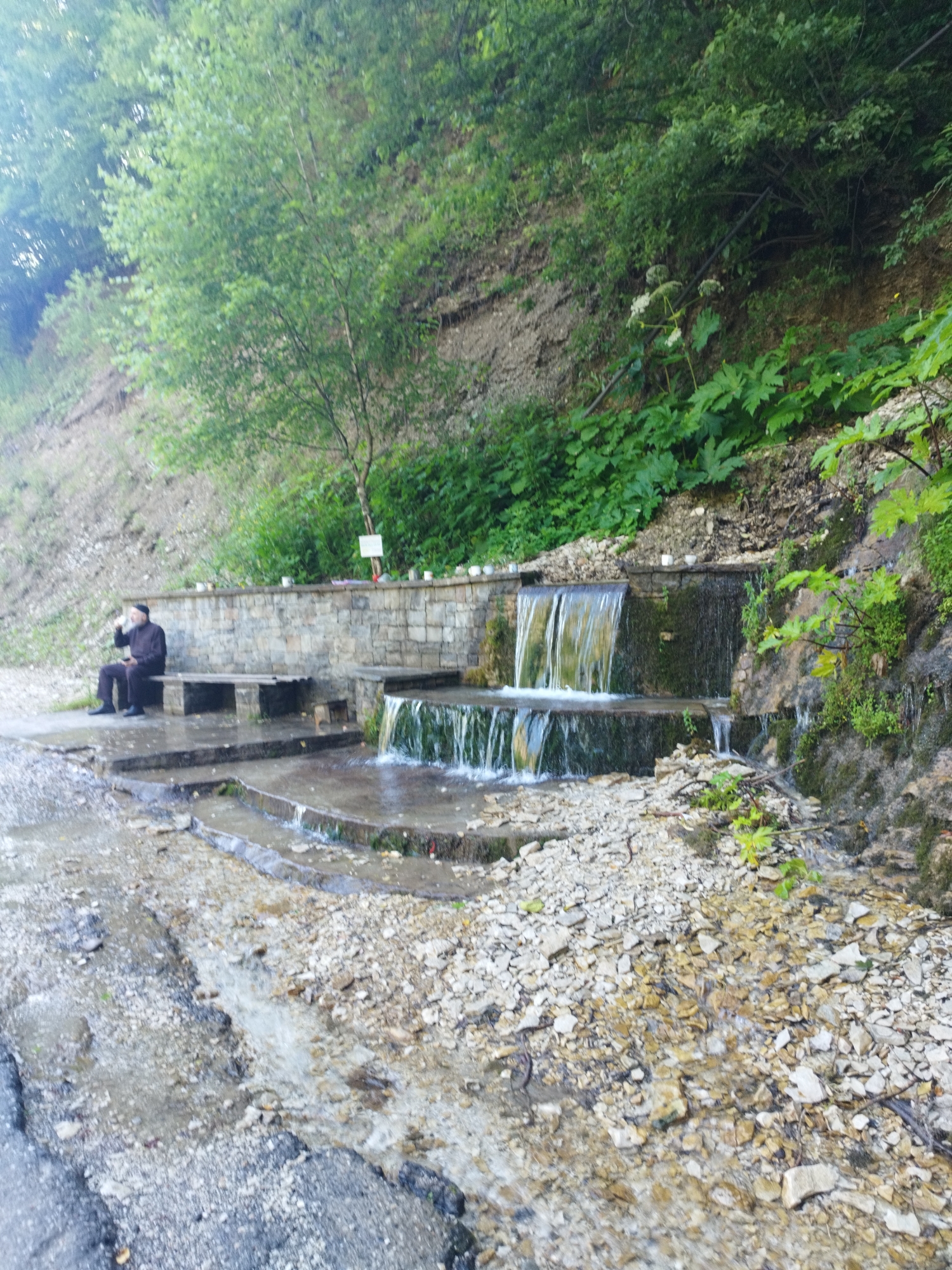 The image depicts a person in dark clothing sitting on a wooden bench next to a tiered waterfall structure. The waterfall is constructed with stone and appears to be flowing over multiple levels into a channel below. The surrounding environment is lush and green, with trees and vegetation on a slope behind the waterfall. There are white cups or containers lined up along the top of the stone wall above the waterfall. The ground in front of the waterfall is covered with stones and some water is flowing across it. The person on the bench is facing towards the waterfall, with one hand raised to their mouth, possibly drinking from a cup. The overall scene suggests a natural setting with some man-made elements.