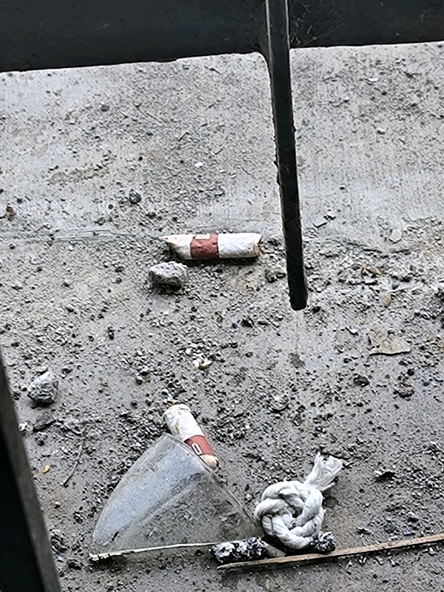 The image shows a close-up, high-angle view of a concrete surface with debris. In the upper half of the image, a dark metal bar runs vertically. Two cigarette butts are visible on the concrete. One is in the upper center of the frame, and the other is in the lower center. Between the two cigarette butts, to the left, is a shattered piece of clear glass, triangular in shape. To the right of the glass, a white rope is tied in a knot. A thin wooden stick is partially visible under the rope. The concrete surface is rough and appears to be littered with small stones and grit. The lighting suggests an outdoor environment with some shadows. The location is unknown.