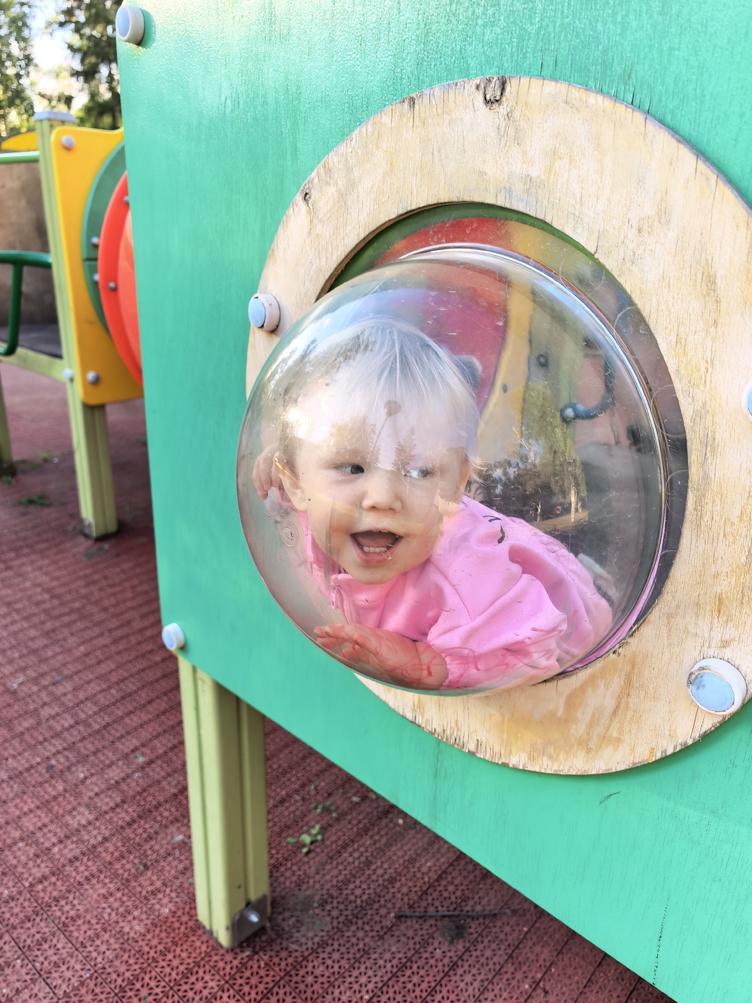 A young child with blonde hair, wearing a pink shirt, is peeking through a clear, dome-shaped window in a green playground structure. The child's mouth is open in a smile. The playground structure has wooden trim around the window and is attached to yellow and red elements, with green supports. The ground is covered with a red, patterned surface. The scene appears to be outdoors, with trees visible in the background. The location is identified as a playground in Sterlitamak, Russia.